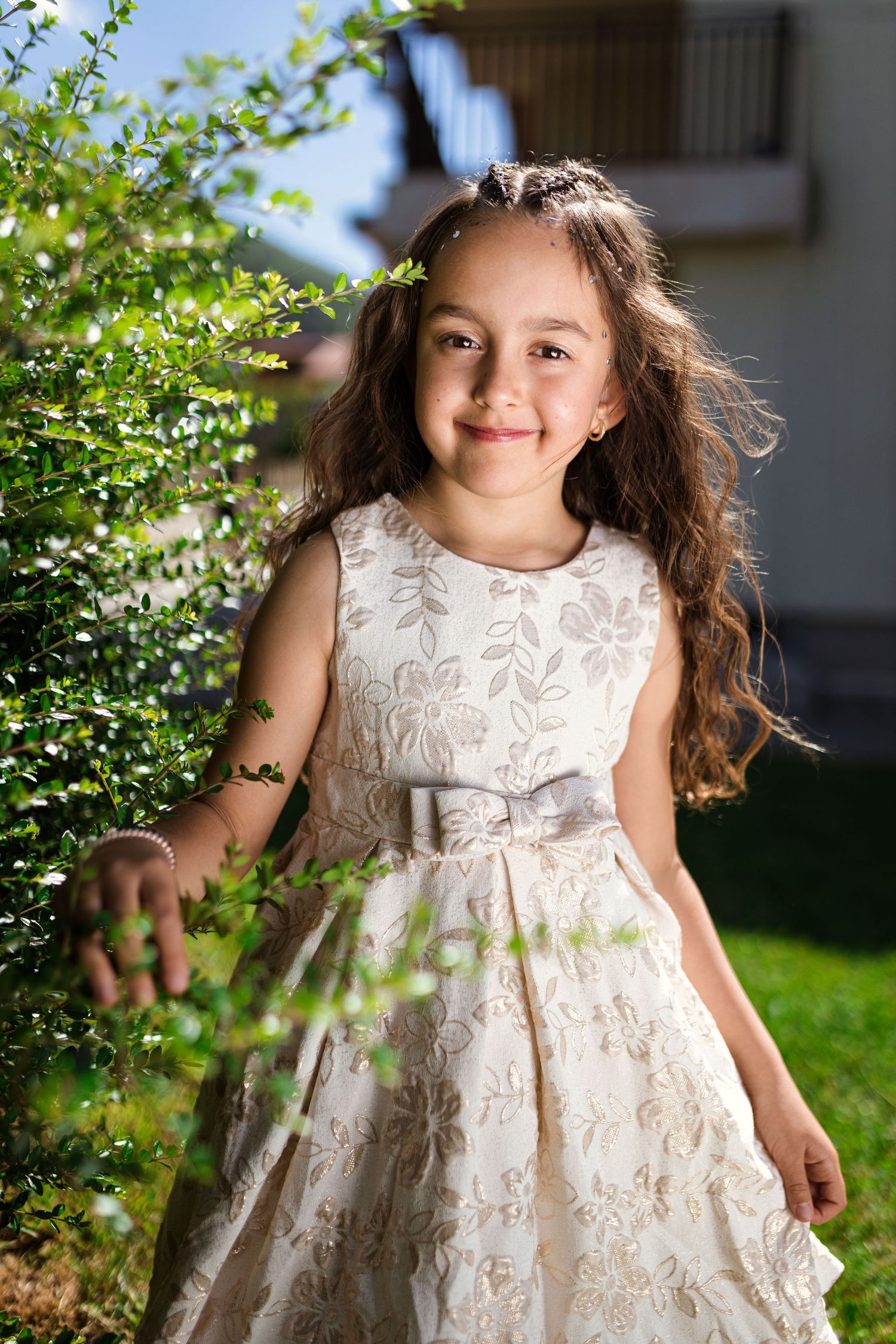 A smiling young girl in a floral white dress stands near green bushes in a sunny garden.