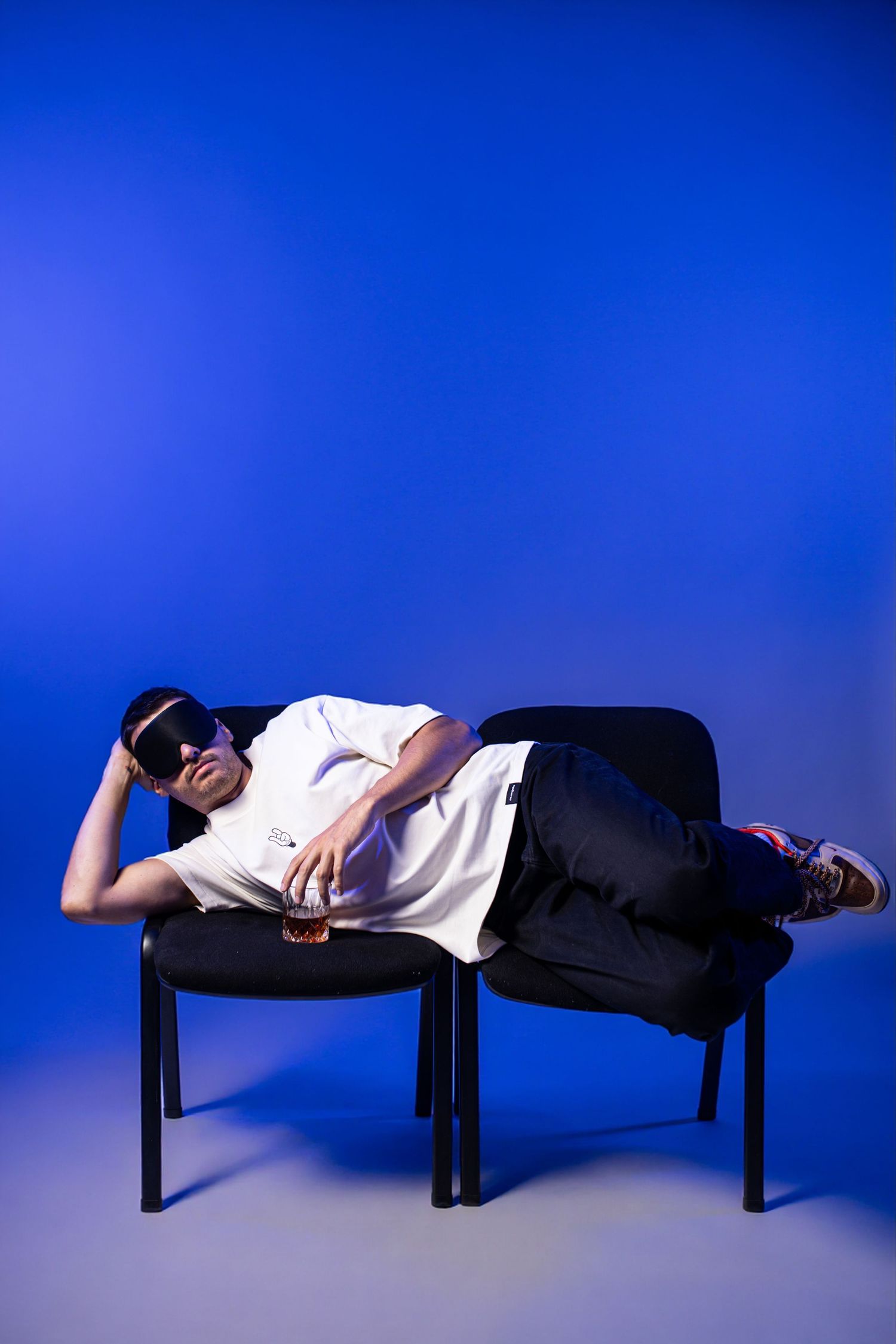 Man in white t-shirt and sunglasses lying across two black chairs holding a drink against a blue background.