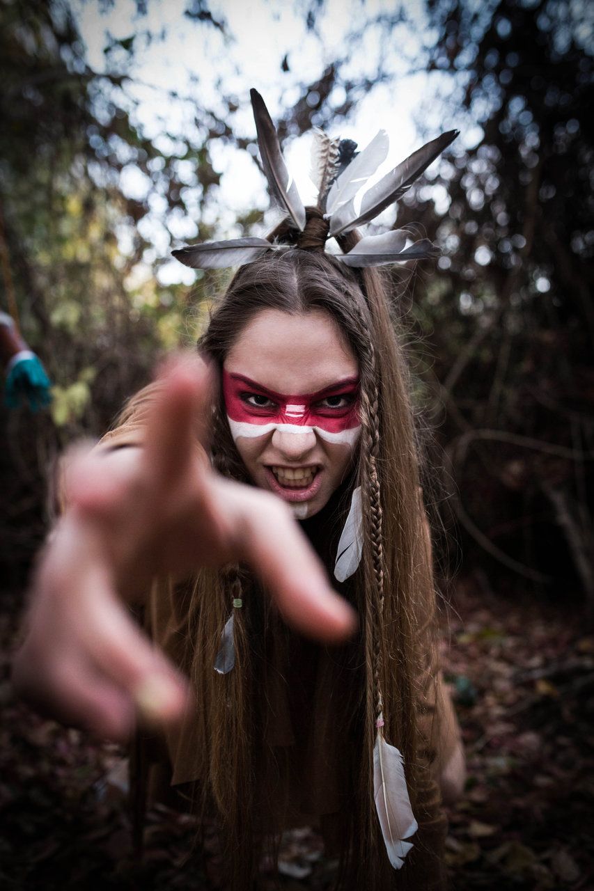 Woman in Native American costume with feather headdress and face paint reaching toward camera in forest.