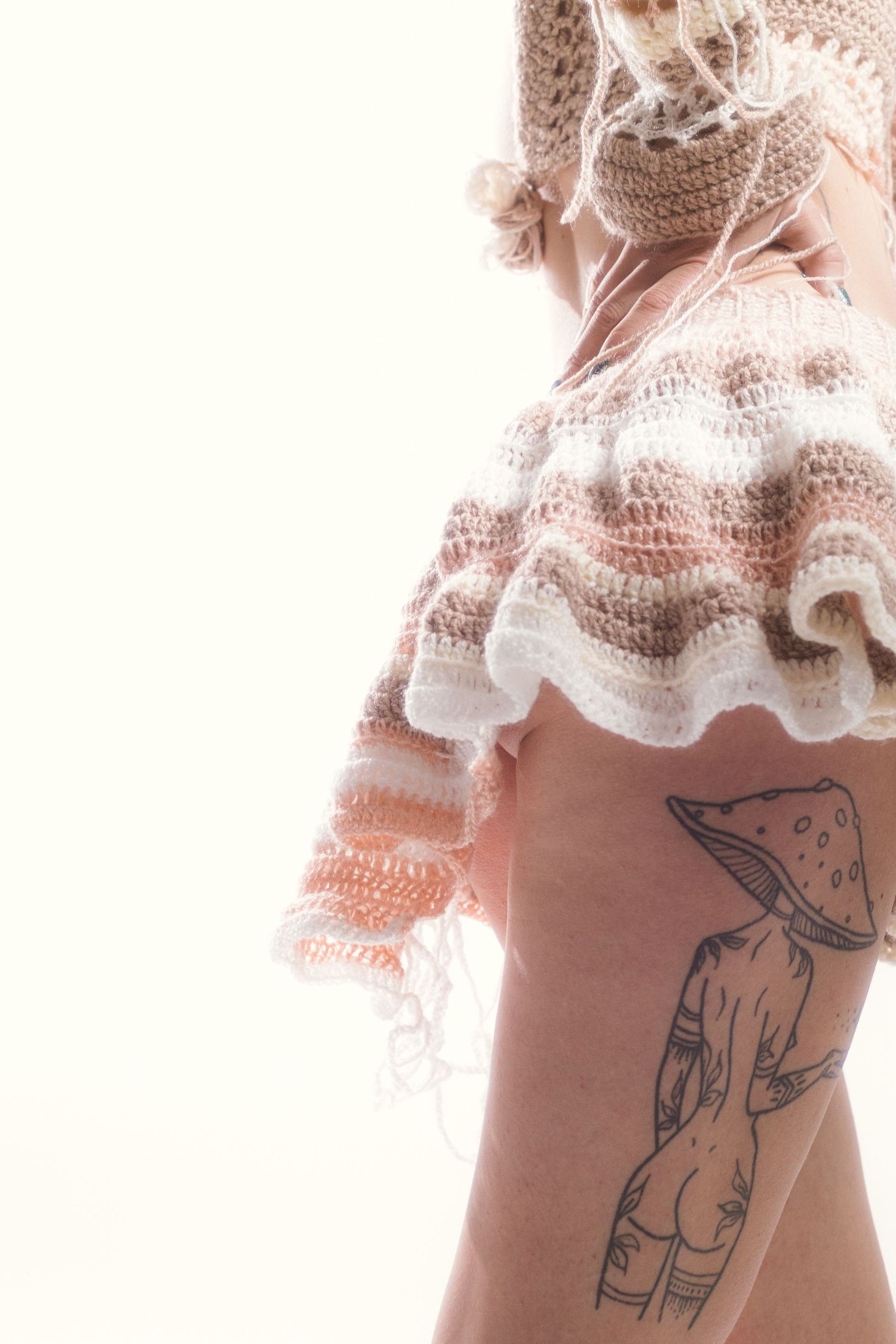 Side view of model in crochet top with mushroom tattoo on hip against white background.