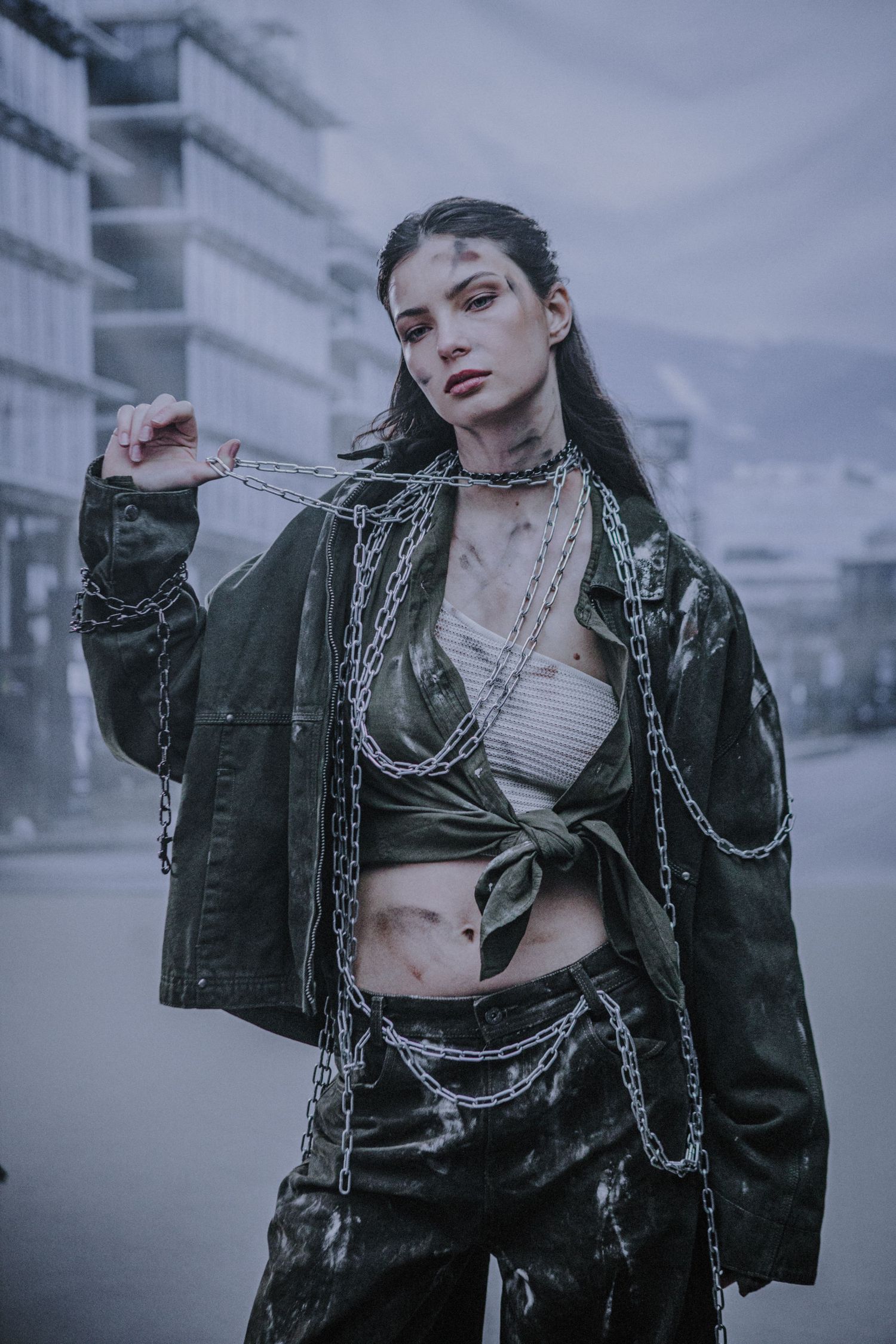 Young woman in olive jacket and chains posing dramatically on a foggy urban street for editorial shoot.