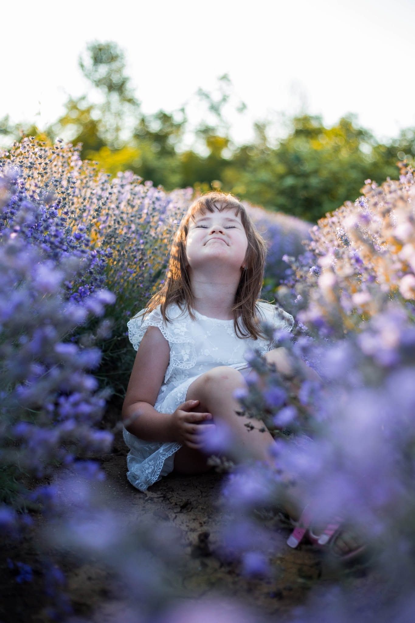 Young girl in white dress sitting joyfully among purple lavender flowers in a blooming field.