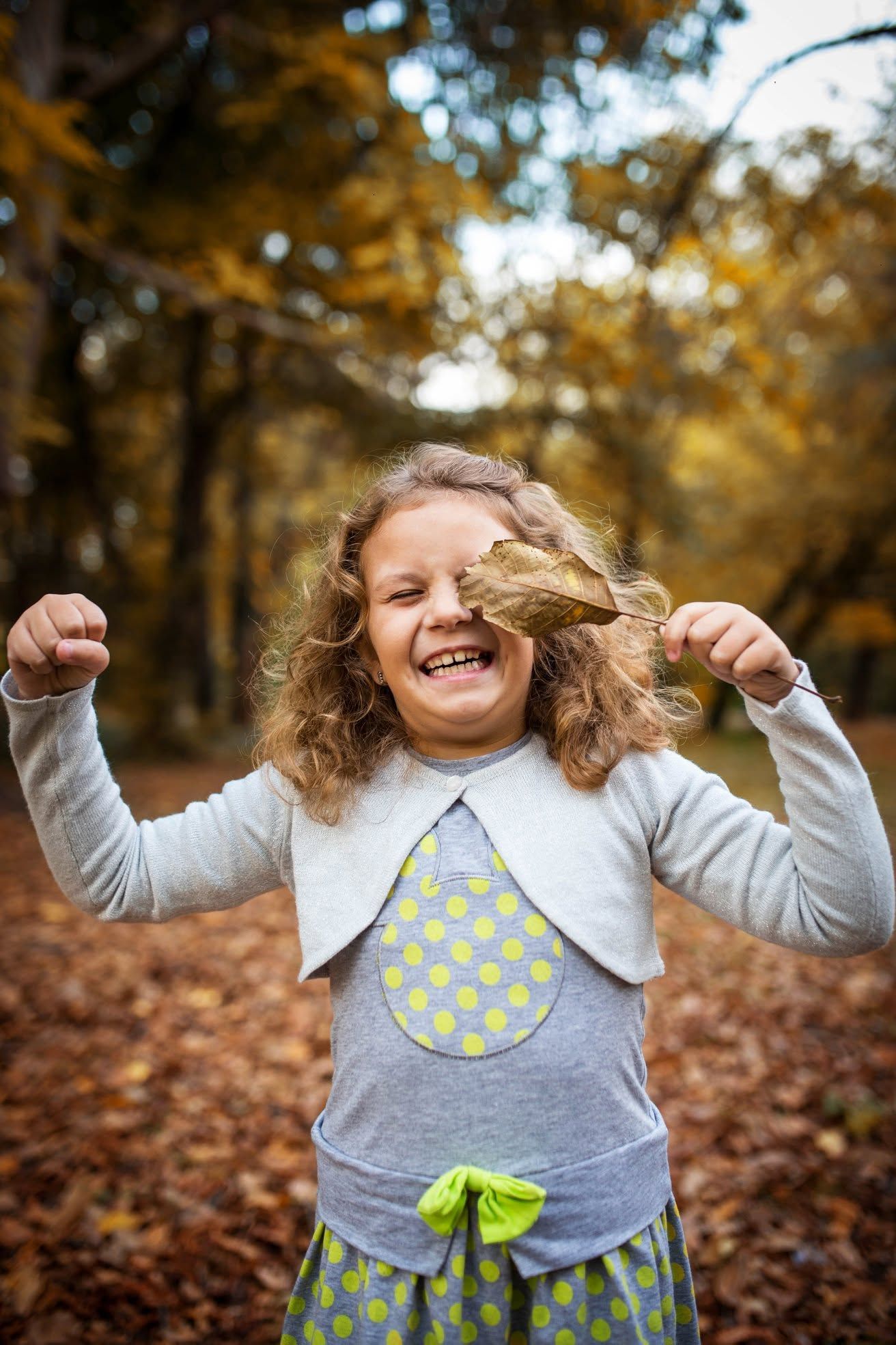 Happy girl laughing in autumn park holding a dried leaf, wearing polka dot dress with curly hair.