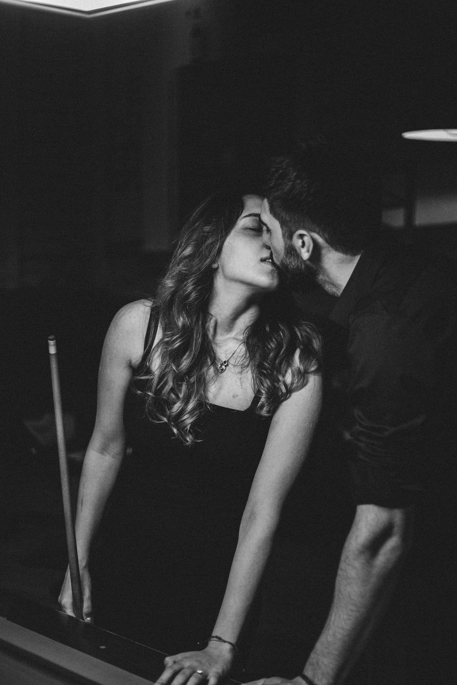 Black and white photo of a couple sharing a romantic kiss near a pool table in a dimly lit bar.