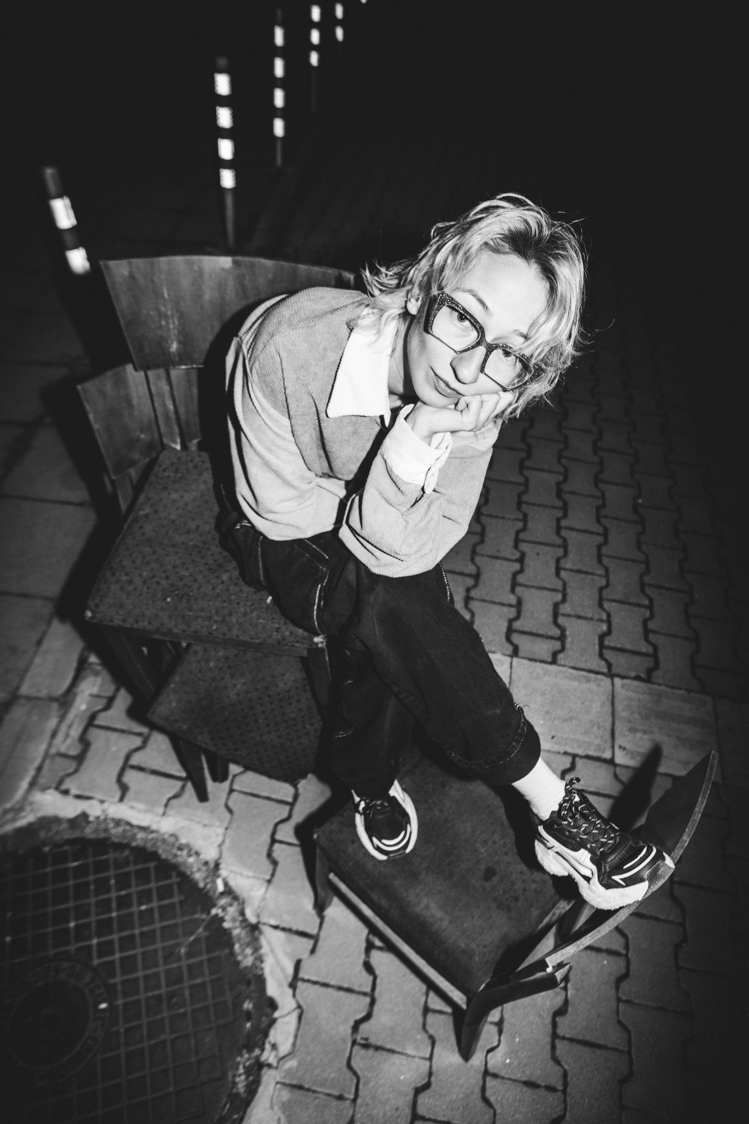 Black and white overhead portrait of a smiling blonde woman with glasses seated on a chair at night.