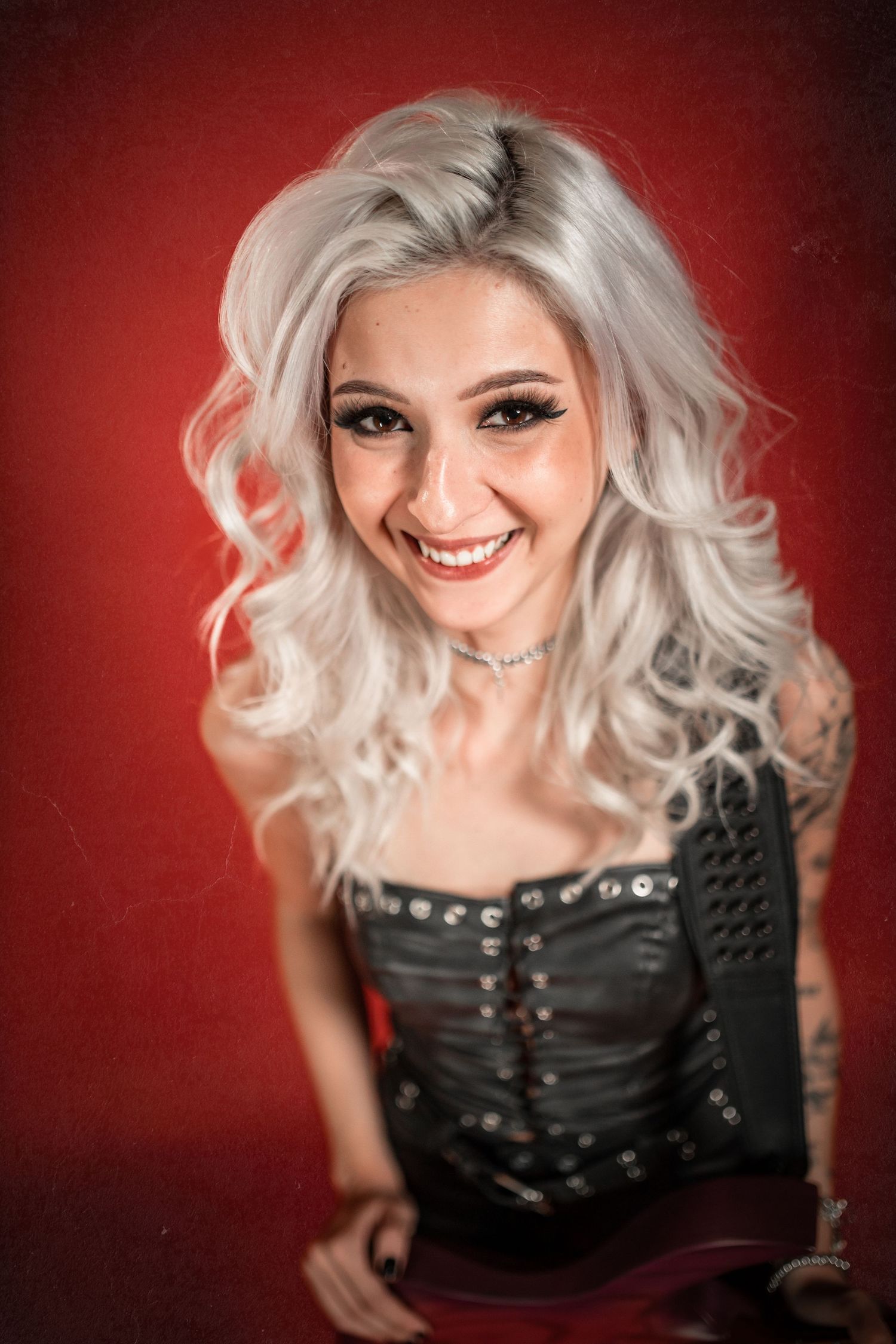 Smiling young woman with silver wavy hair wearing black studded corset against red background.