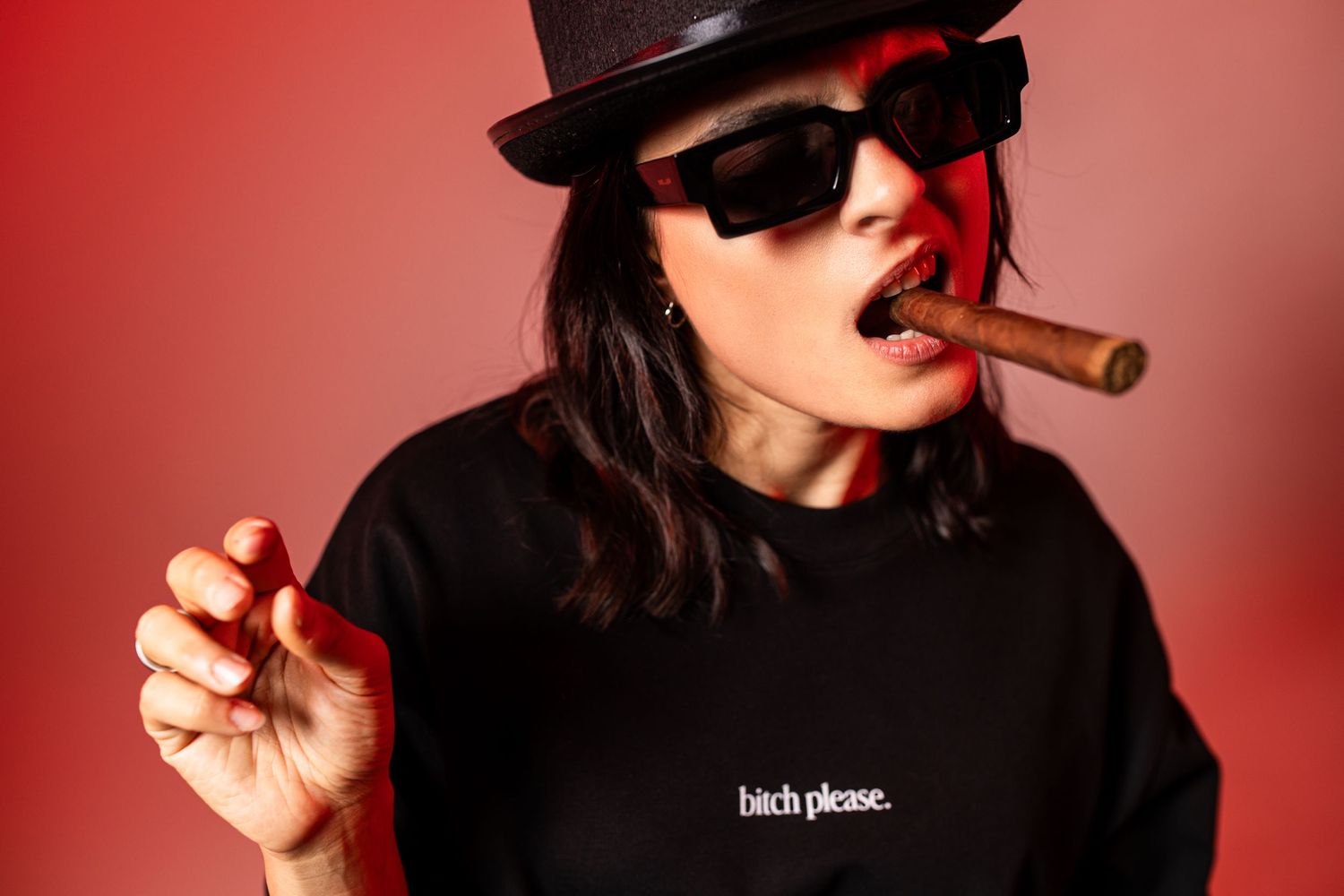 Woman in black hat and sunglasses smoking a cigar, wearing a black sweatshirt with bold text on red background.