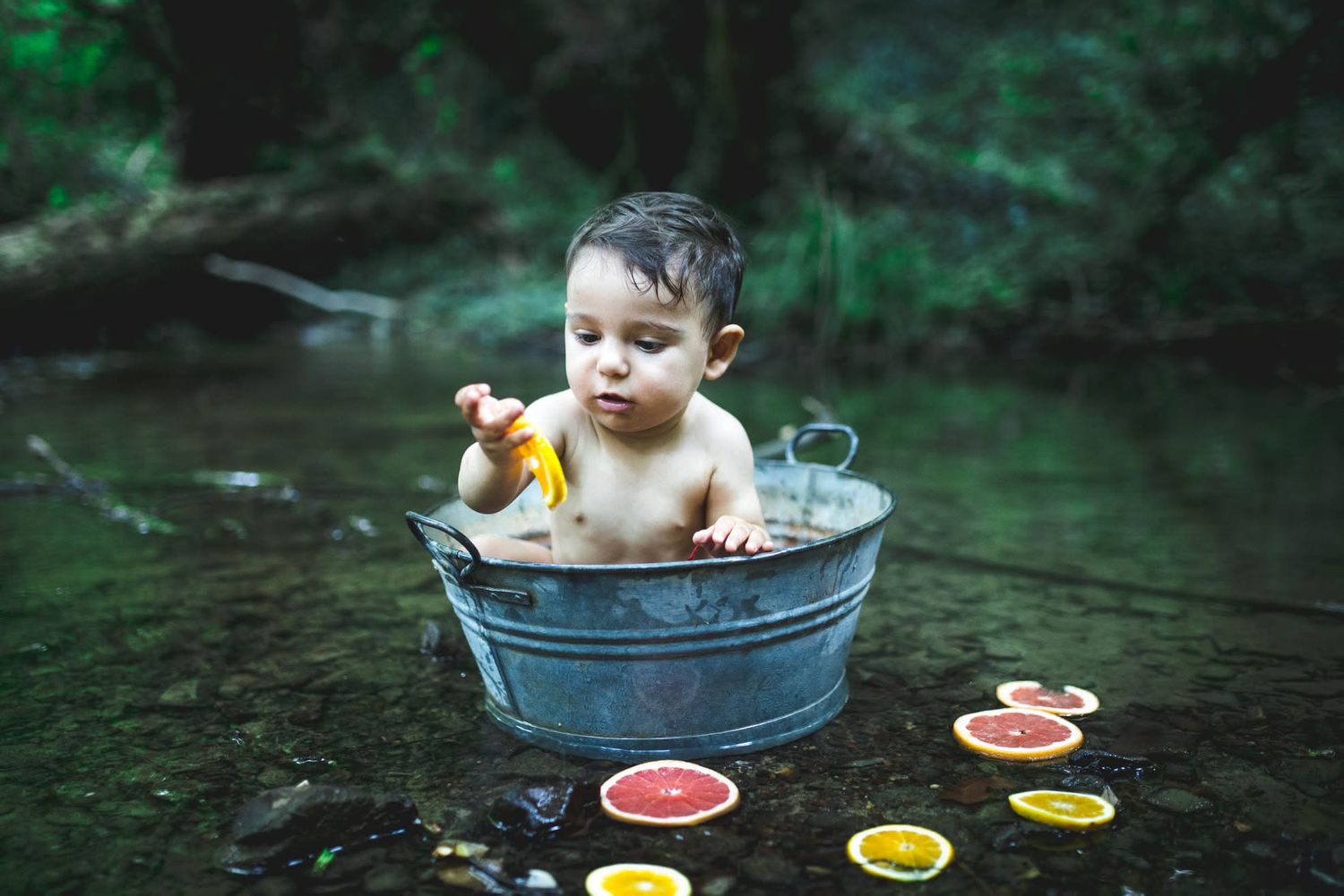 A baby sits in a metal tub outdoors in a stream, playing with citrus fruit slices floating in the water.