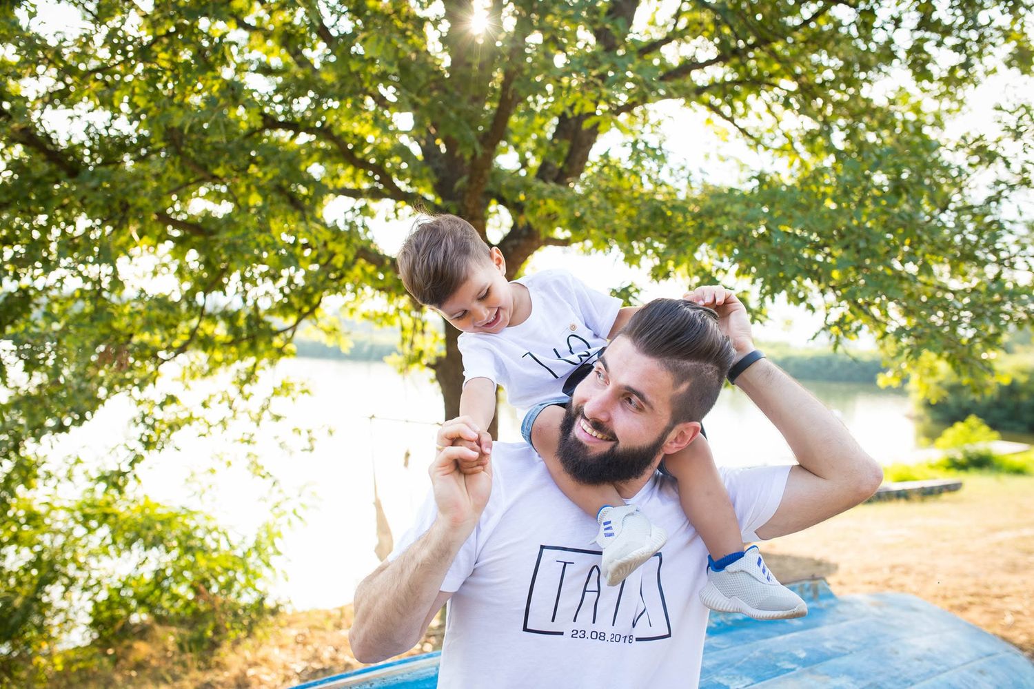 A bearded father carries his young son on his shoulders outdoors near a lake, both wearing matching white t-shirts.