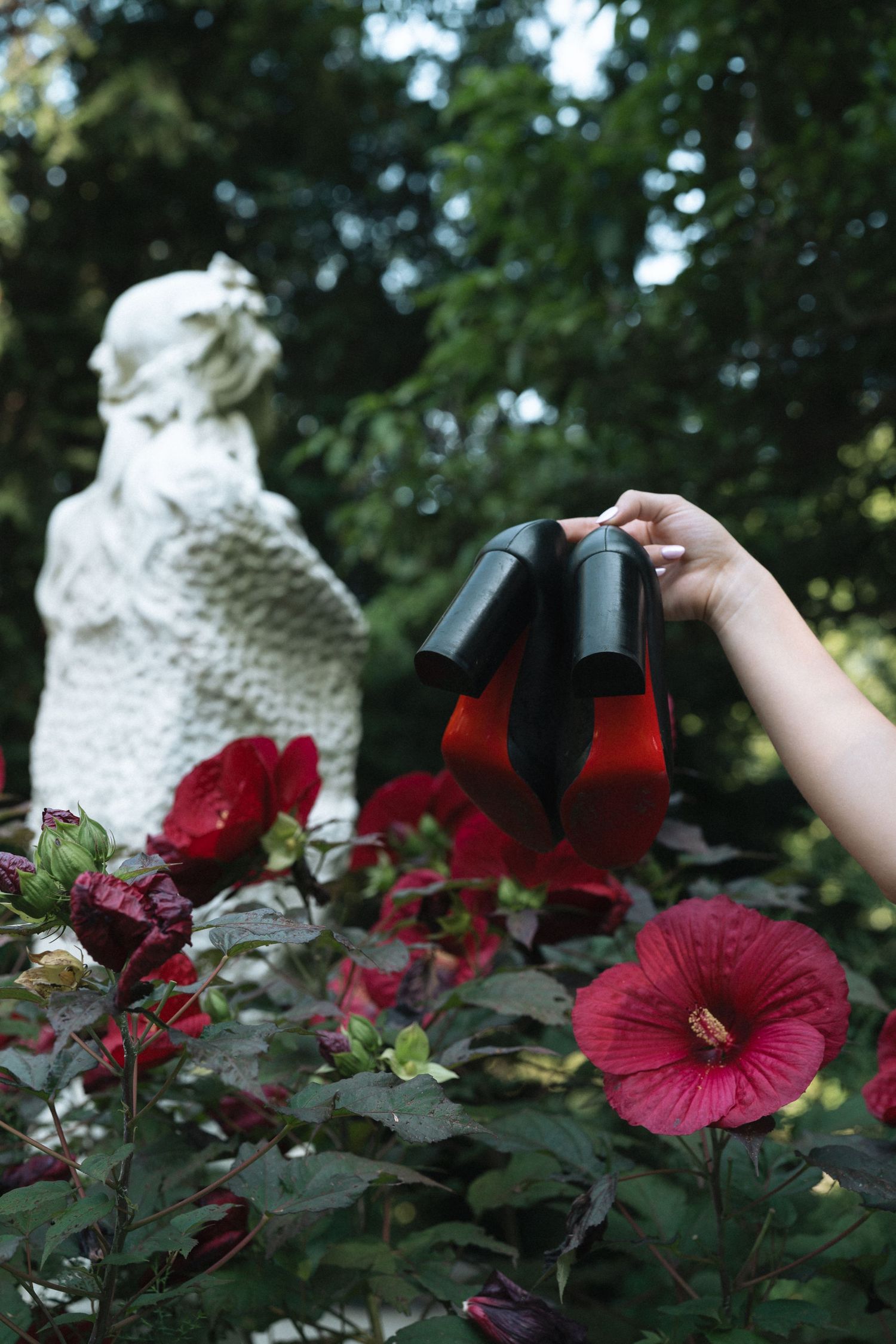 A hand holds designer shoes over vibrant red hibiscus flowers with a white statue in the garden background.