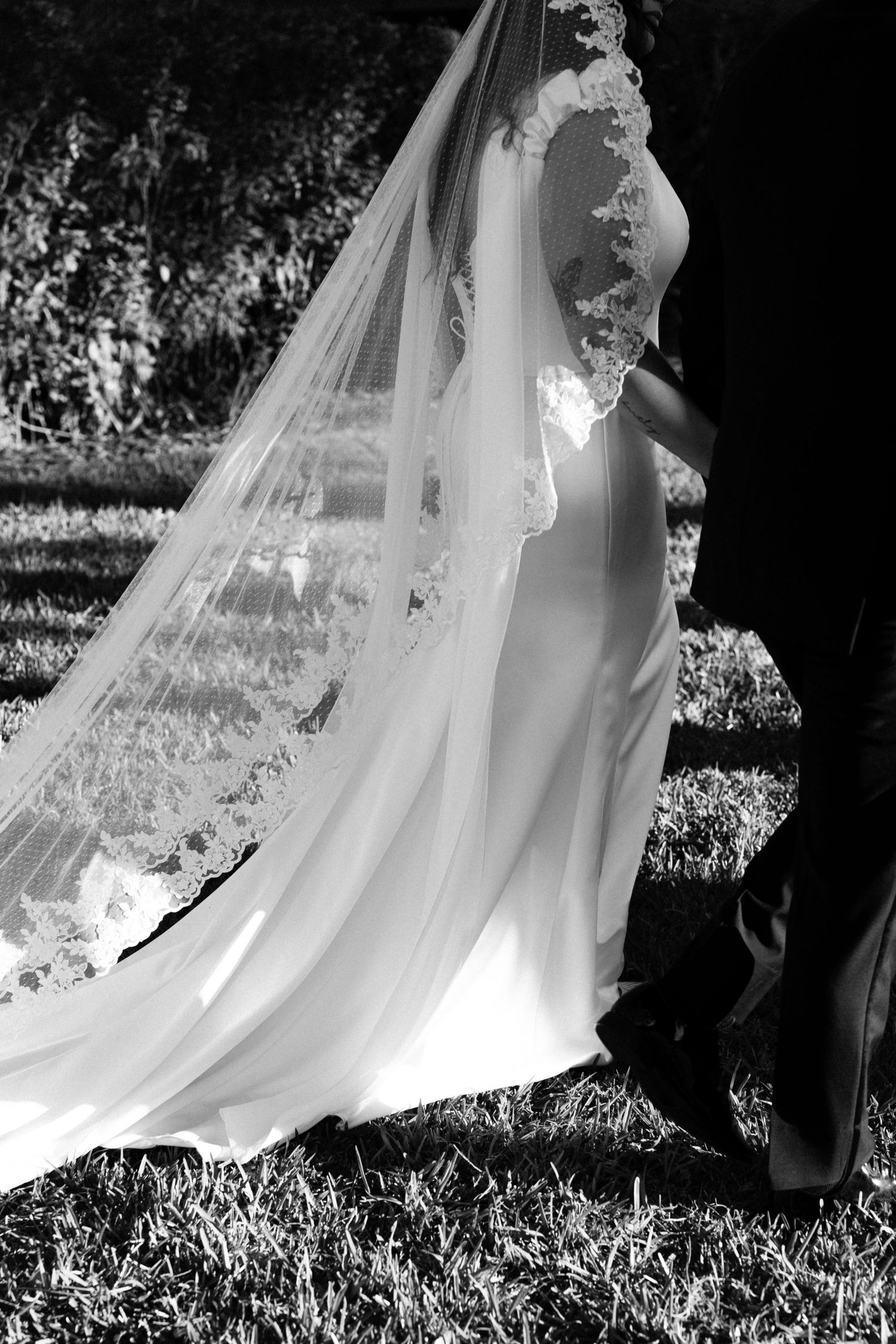 A flowing white wedding veil catches sunlight and creates an ethereal silhouette in a black and white outdoor photograph.