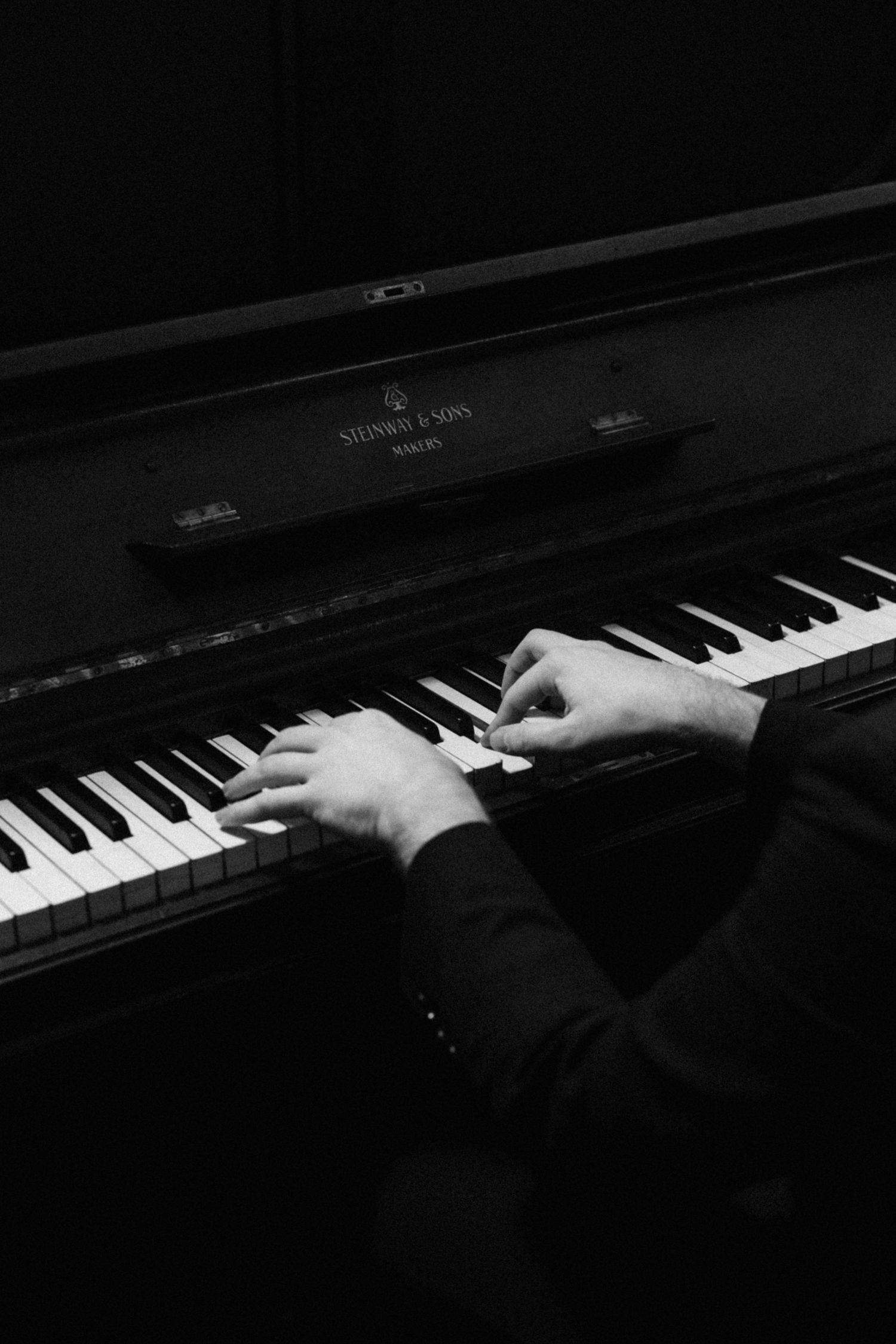 Hands play piano keys on a black grand piano in a dramatic low-key black and white composition.