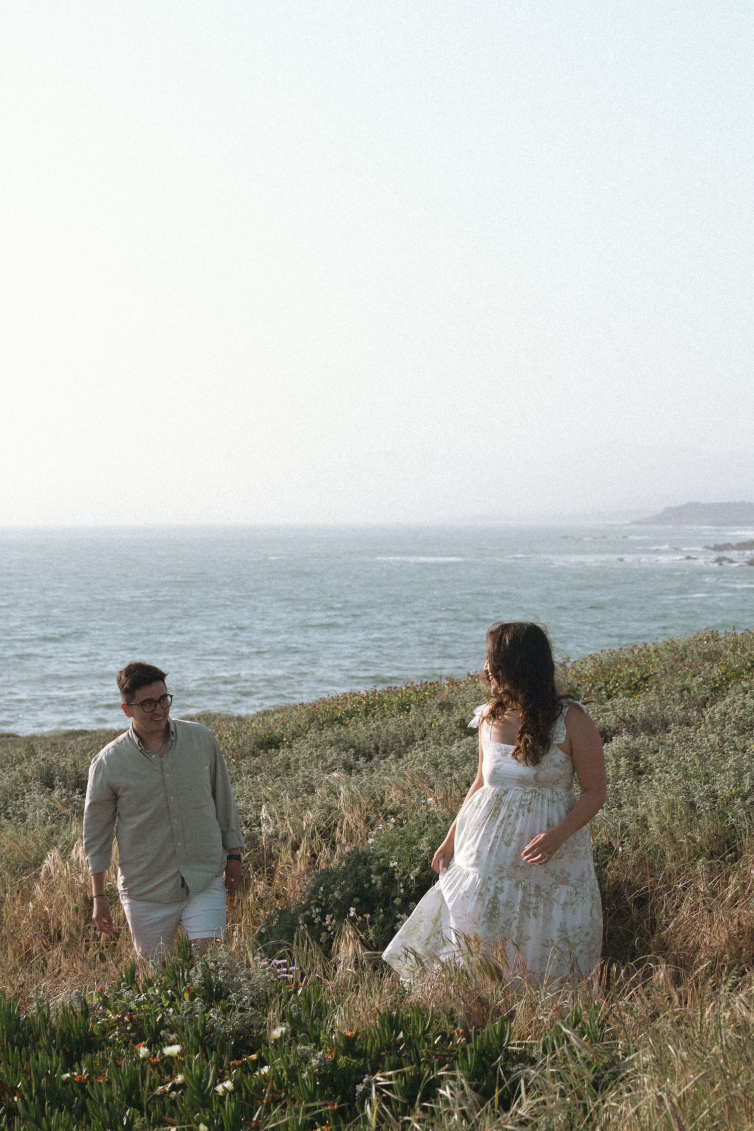 A couple  walks along a California coastal cliffside with Pacific Ocean views and wildflowers on a misty afternoon.