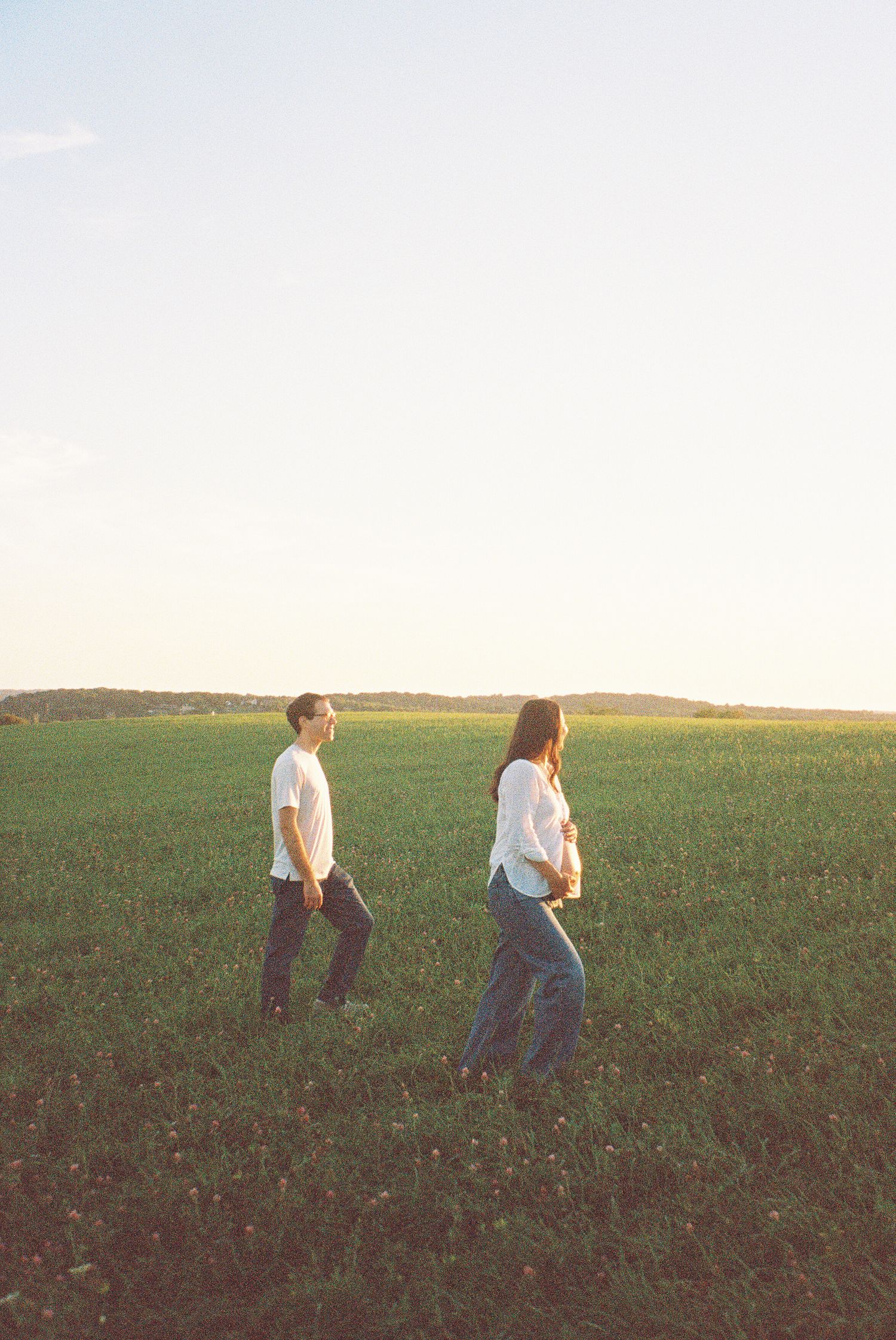 A couple in white shirts and jeans walk through a lush green field at golden hour sunset captured on vintage film.