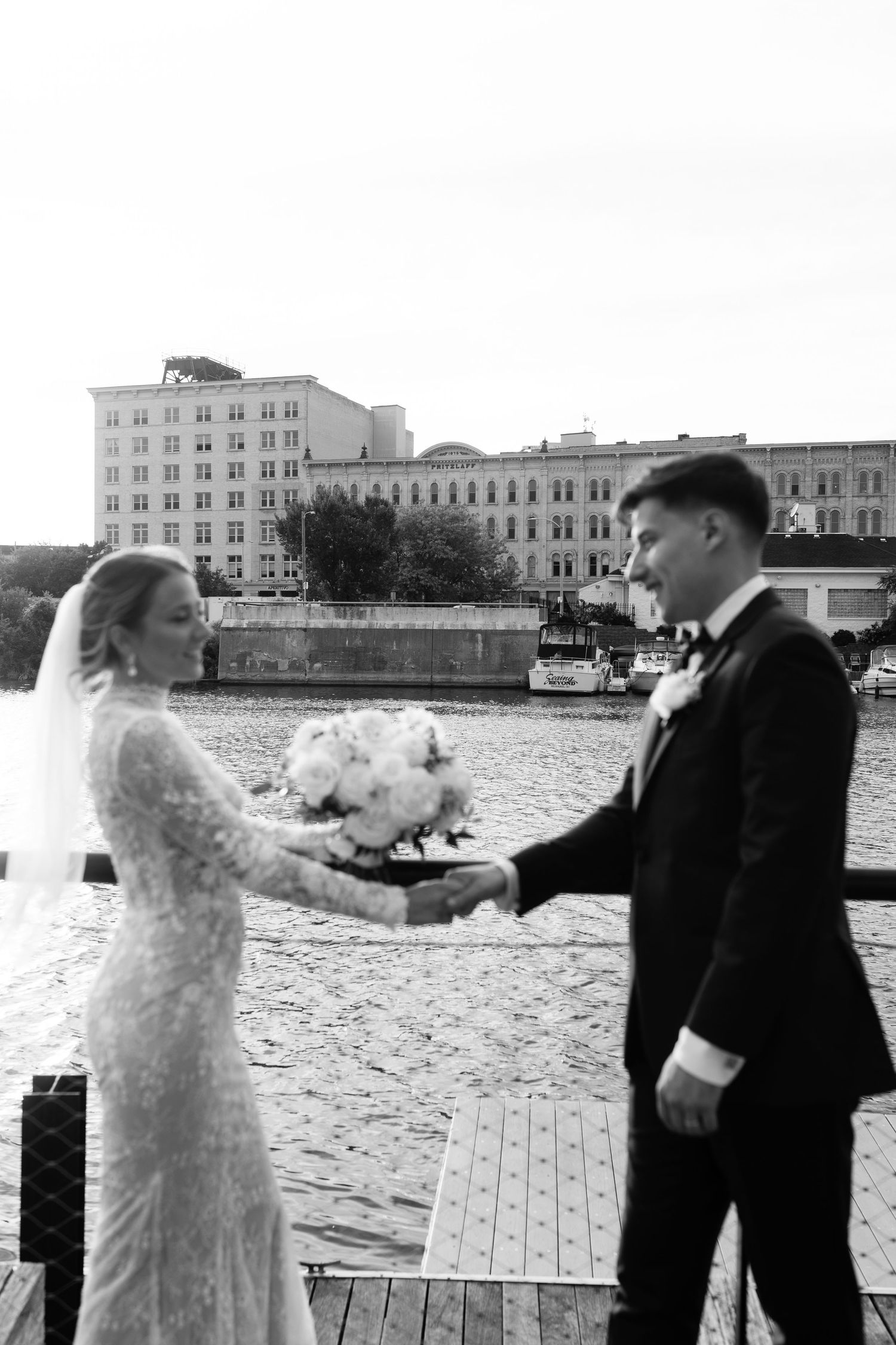 Black and white photo of bride and groom holding hands on a waterfront dock with historic buildings in the background.