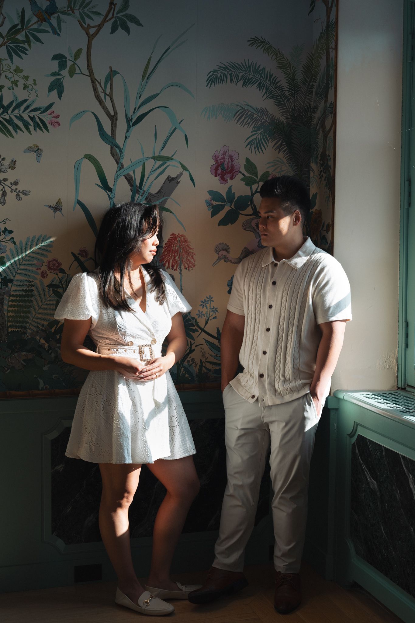 A couple in light-colored outfits stands intimately against a wall with floral wallpaper and palm frond designs.
