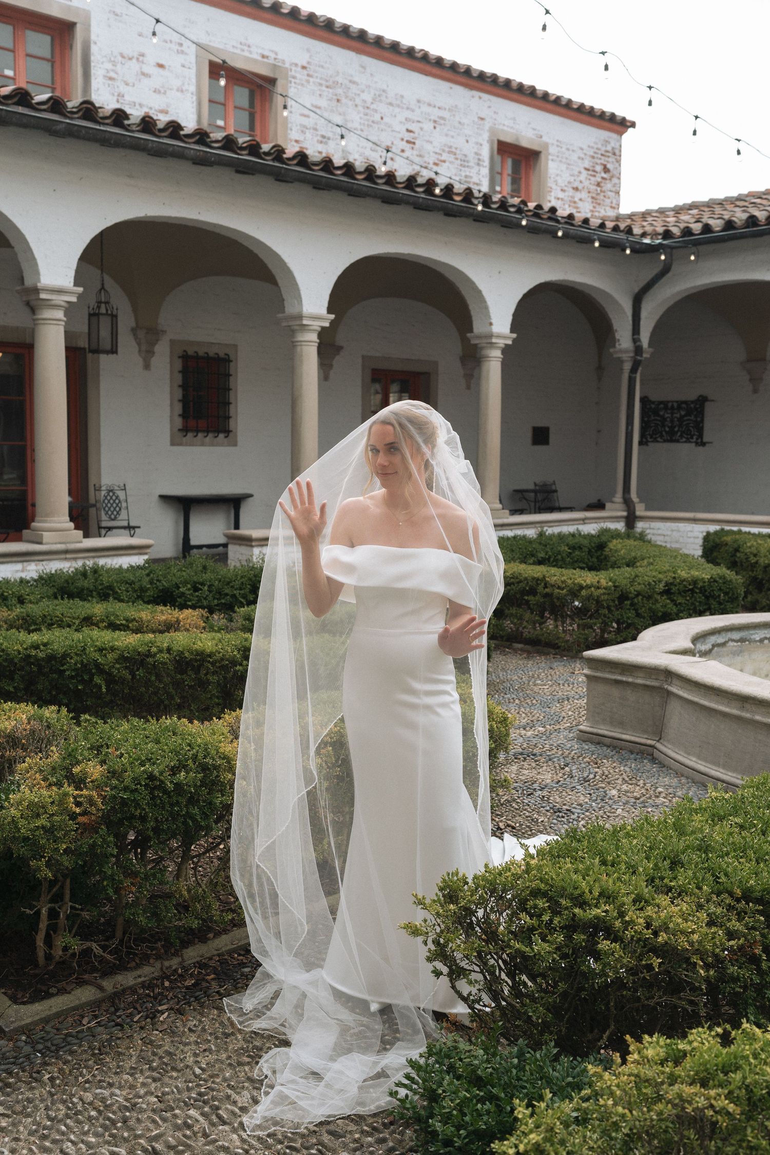 European bride in white off-shoulder gown and flowing veil poses in elegant Italian courtyard garden with arched colonnade.