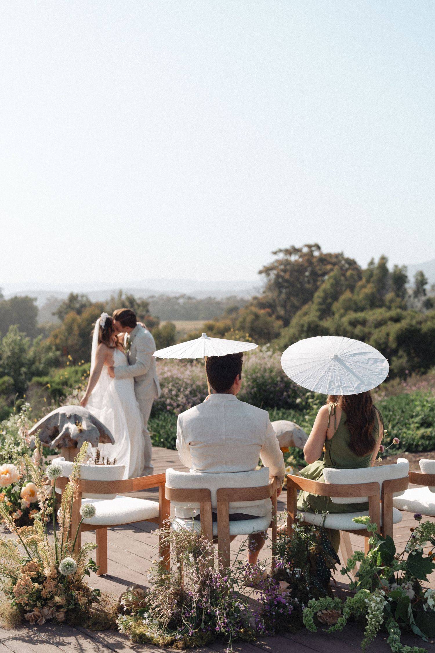 Wedding guests sit in white chairs surrounded by lush florals and greenery at an outdoor ceremony overlooking rolling hills in Santa Barbara