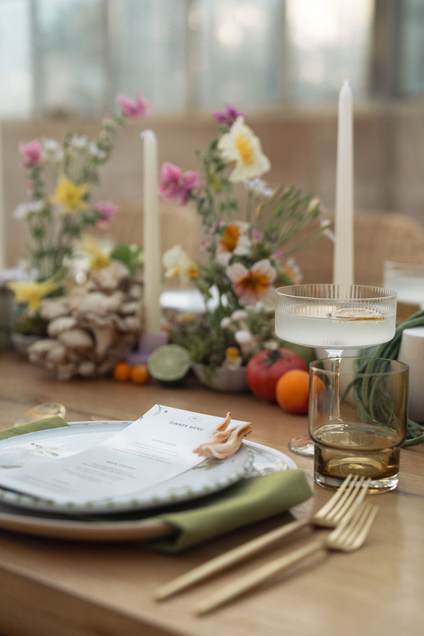 Elegant table setting with gold cutlery, green napkin, menu card, candles, and colorful floral centerpiece on wooden table in Santa Barbara