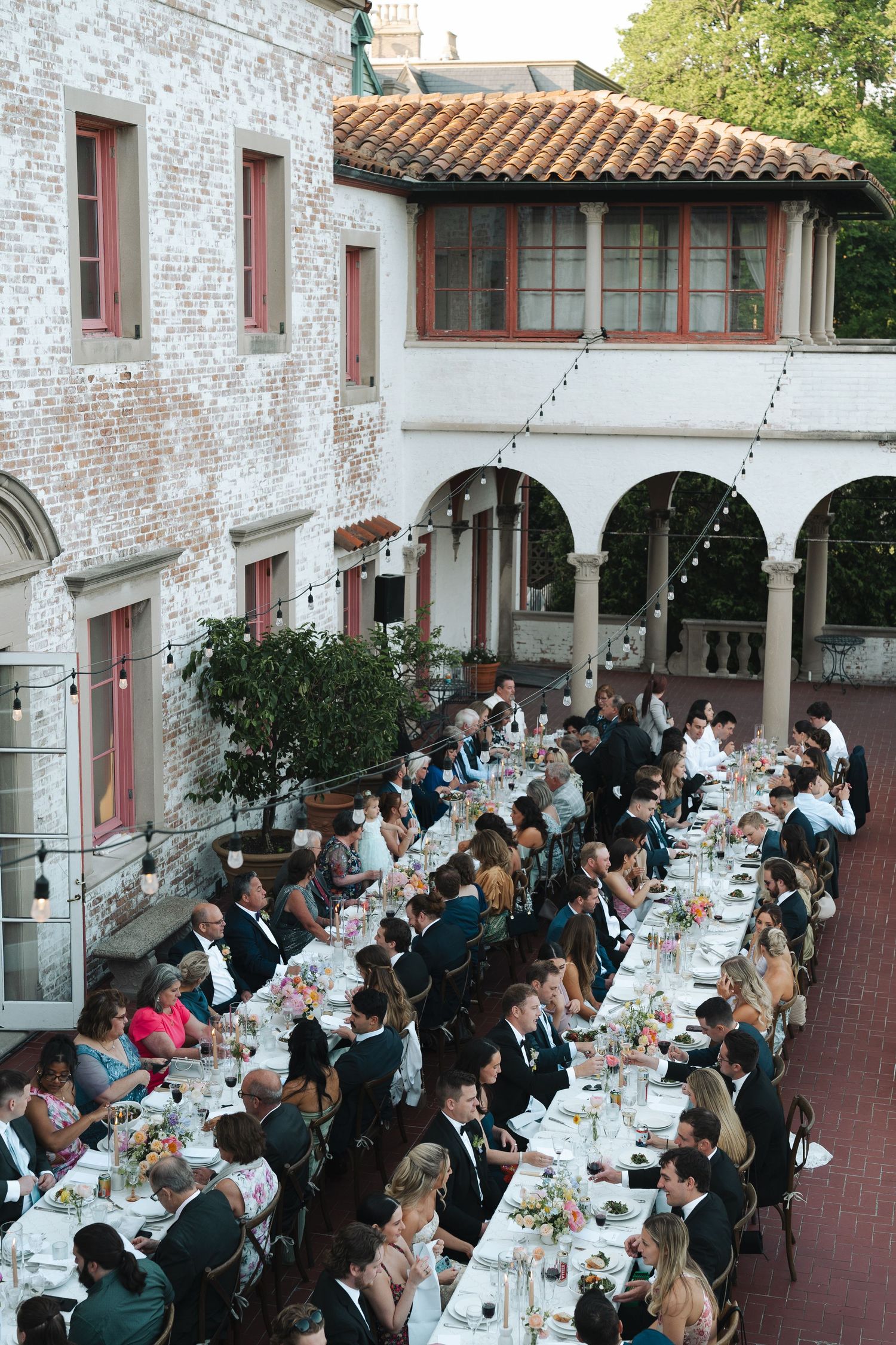 Large formal outdoor dining reception with guests seated at long tables under Italian-style arched architecture and brick walls.