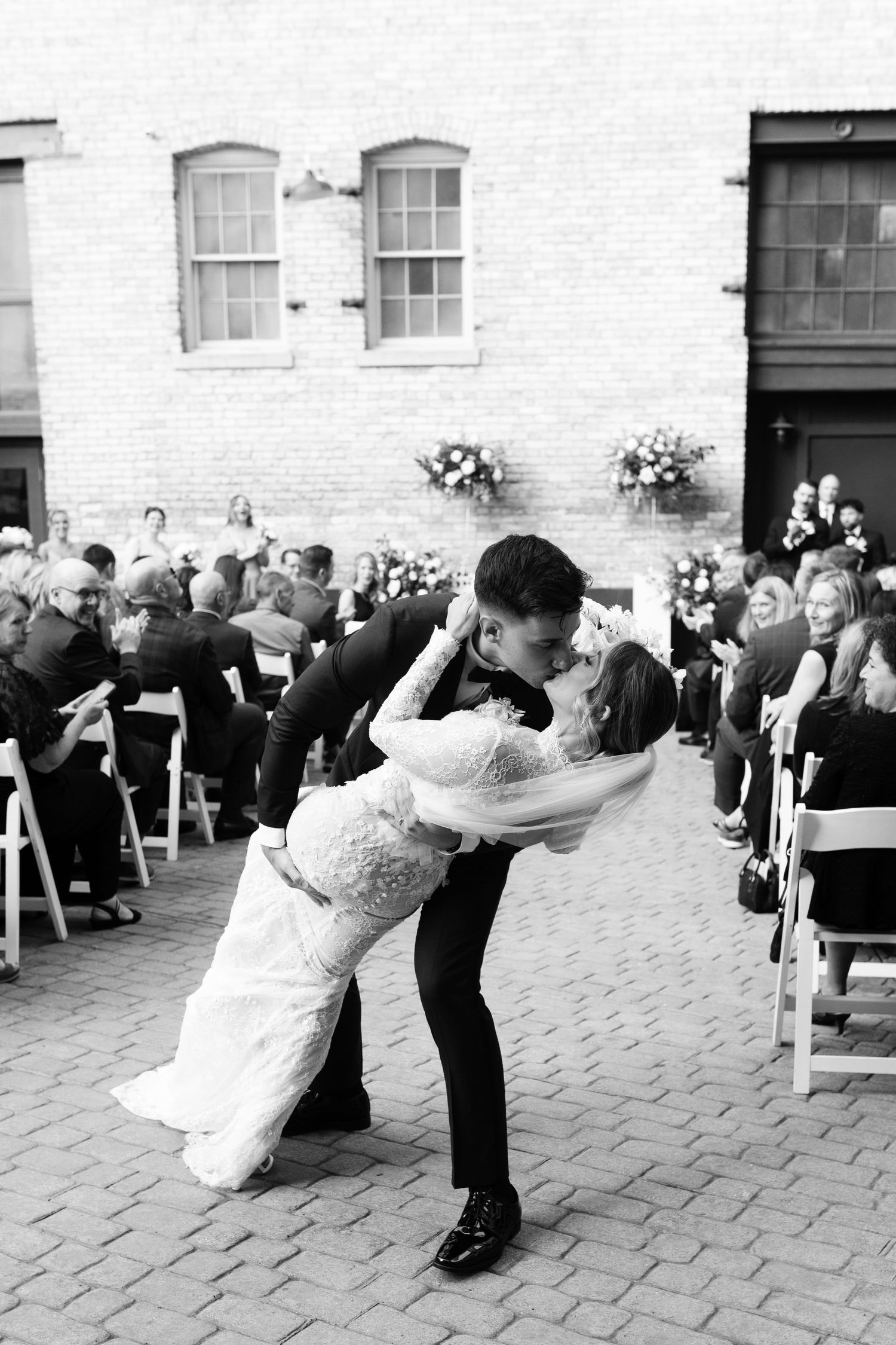 A romantic dip kiss during a wedding ceremony on a brick patio with white chairs and floral decorations in black and white.