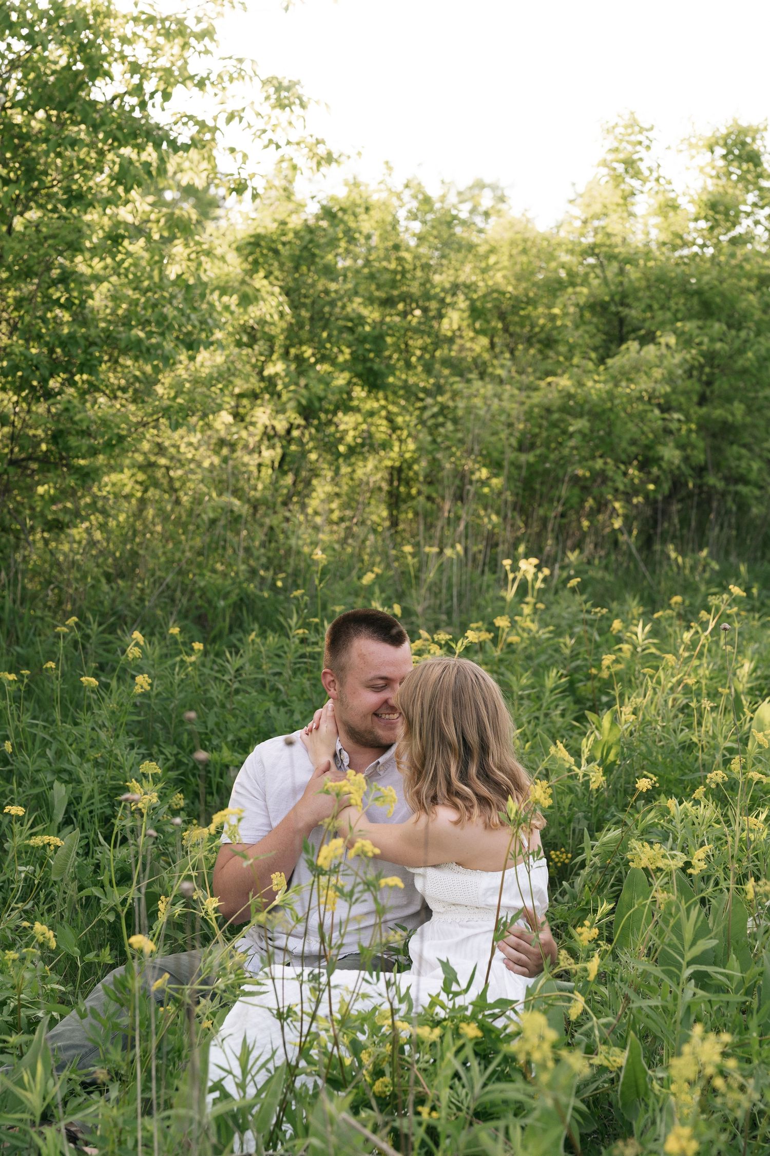 A couple sits together in a lush green meadow with yellow wildflowers, sharing a tender romantic moment outdoors.