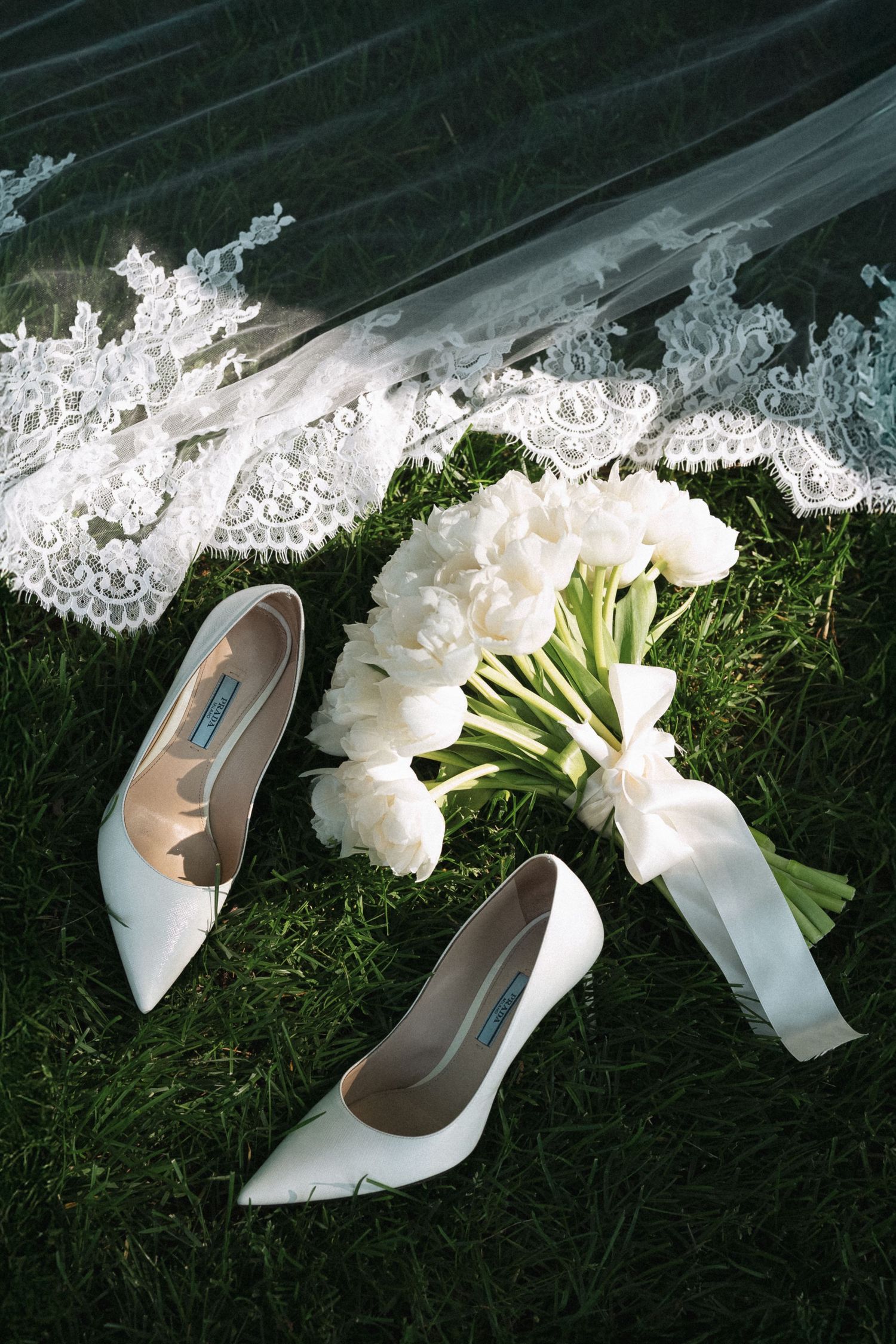 White wedding Prada high heels and bridal bouquet of white tulips arranged with delicate lace veil on grass.