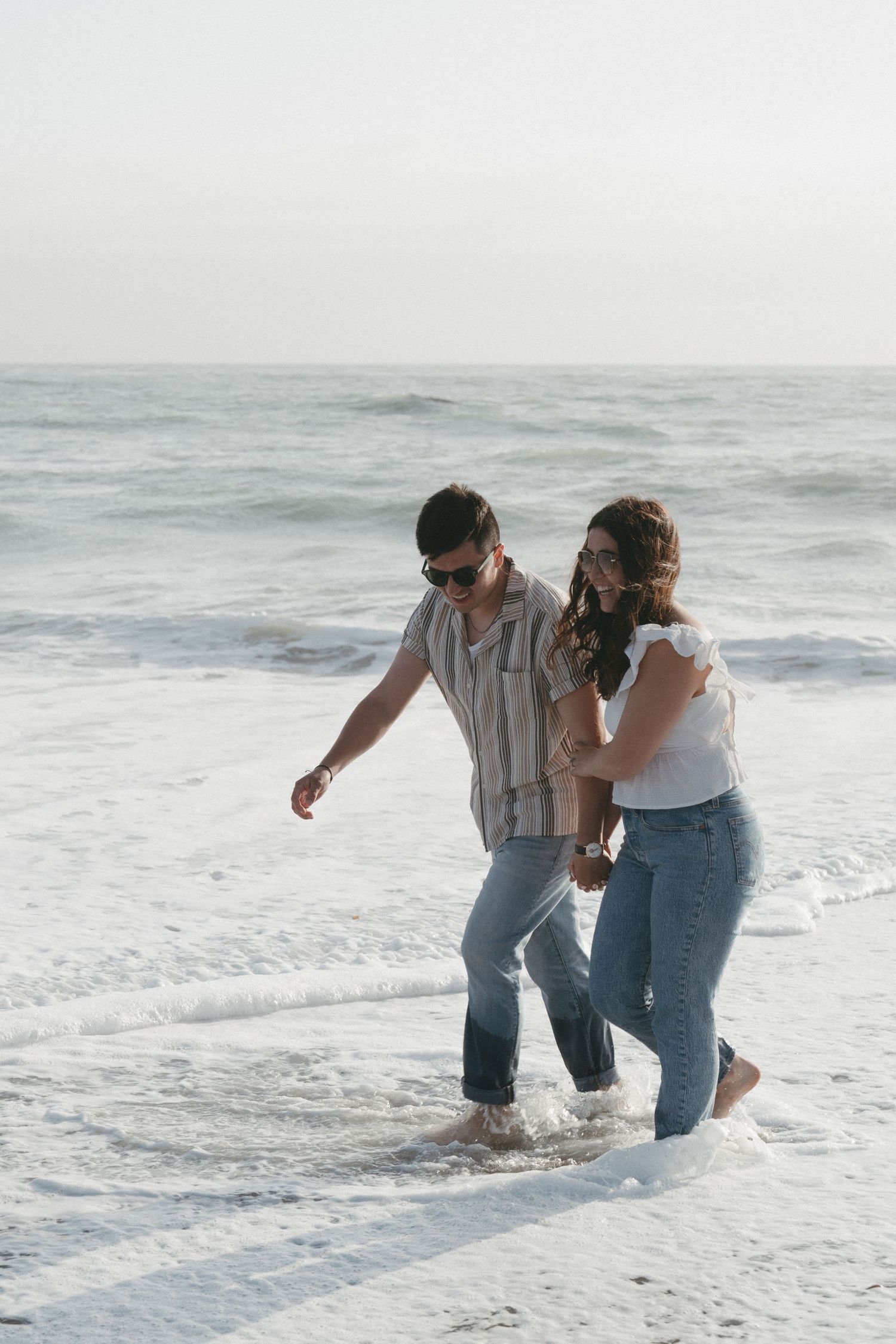 Couple walking hand in hand through shallow ocean waves on a misty beach in California