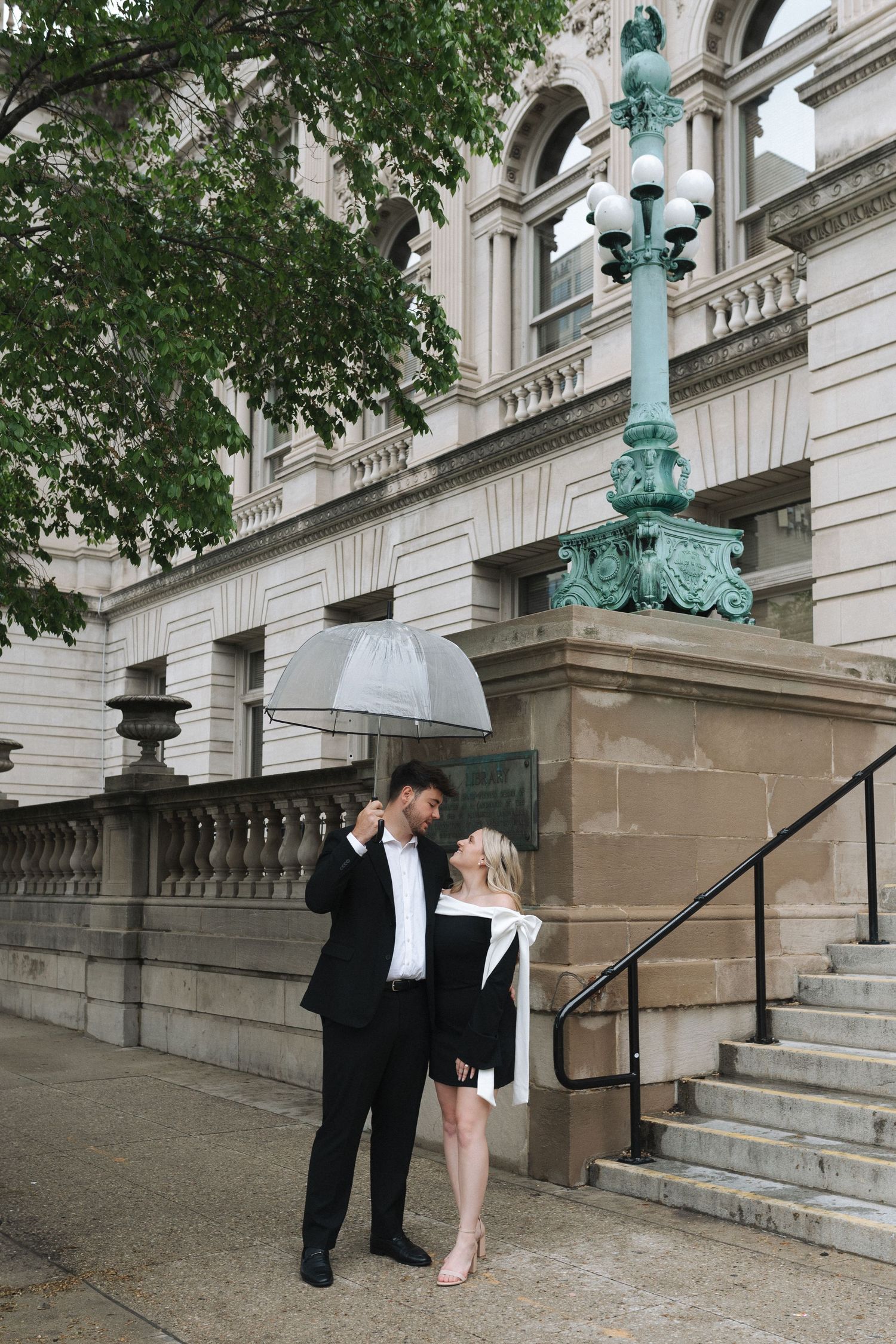 Couple sharing umbrella in rain outside classic stone building with ornate green lamp post.