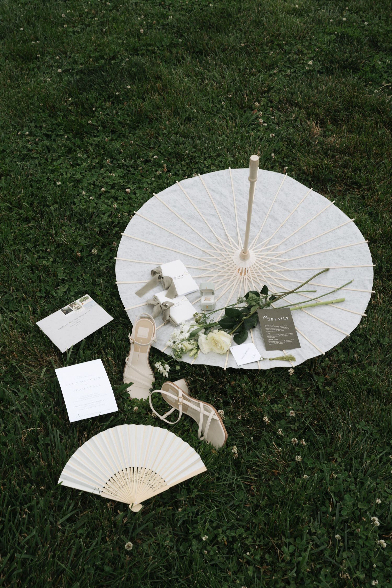 White parasol, hand fan, roses, sandals, and wedding stationery arranged on green grass for a bridal flat lay.