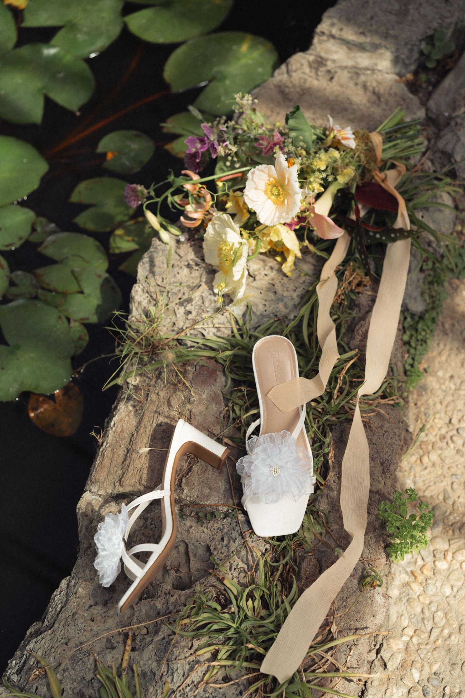 White floral shoes next to beige ribbons adorn a stone surface against a backdrop of green foliage.
