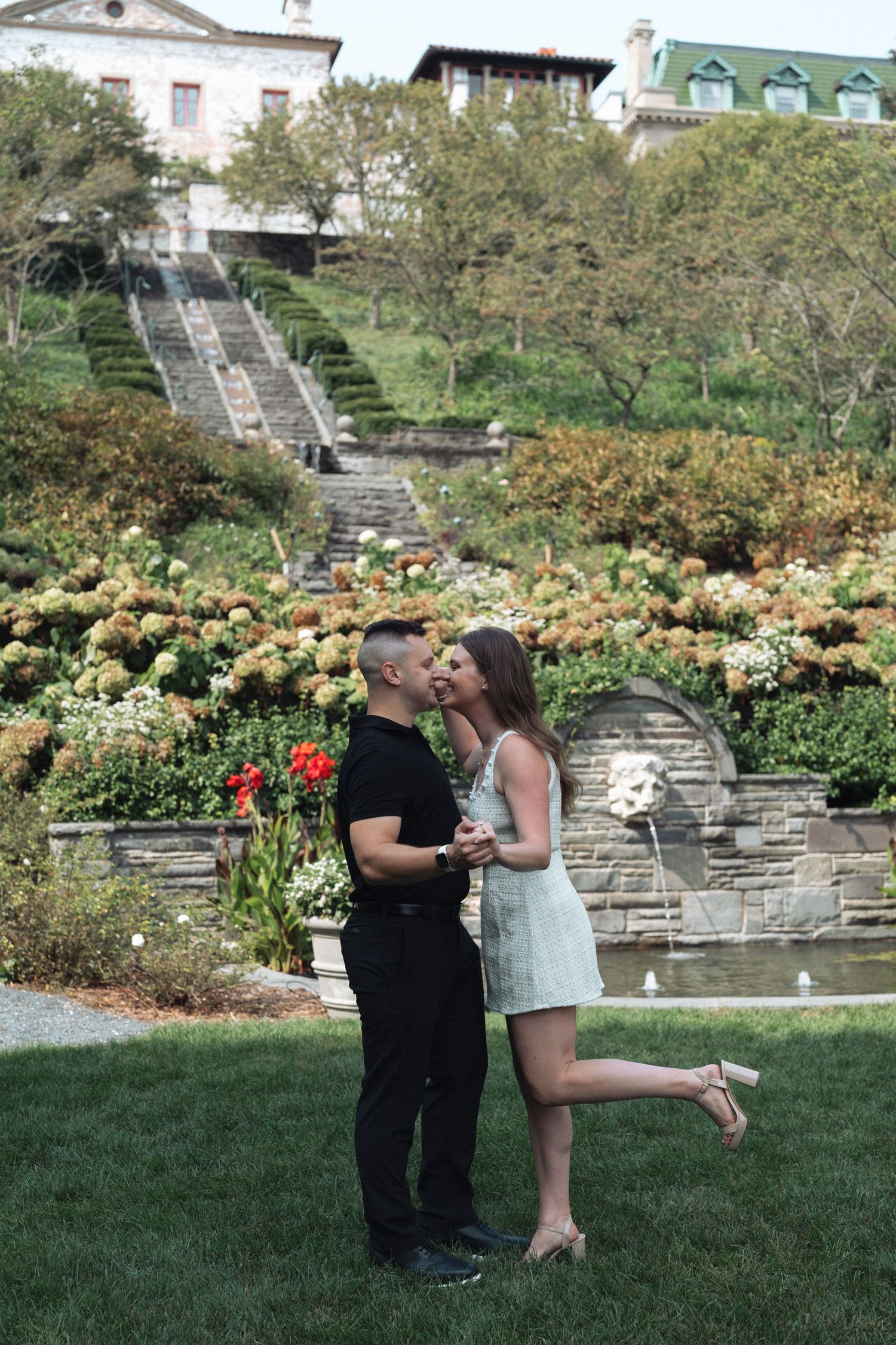 Couple sharing a romantic kiss in a beautiful garden with stone stairs, blooming flowers, and an elegant European estate in the background.