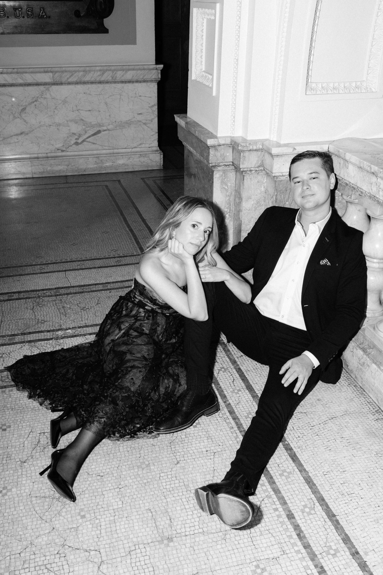 Black and white old money style photo of elegant couple sitting on marble steps, woman in lace gown, man in suit.