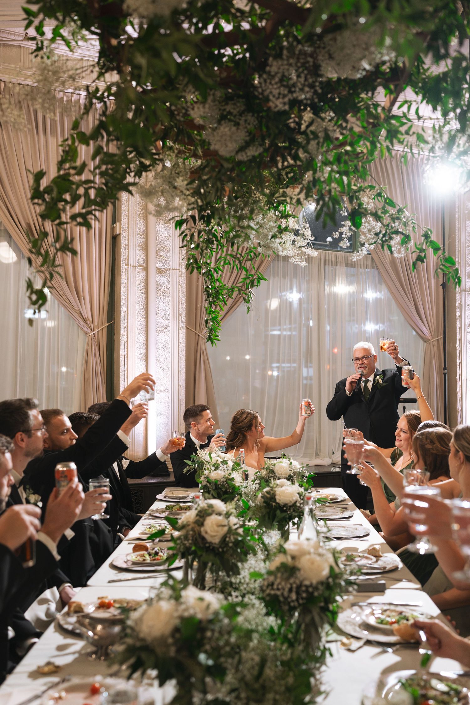 Wedding guests raise glasses in a toast at a long reception table with white floral centerpieces and hanging greenery in an elegant venue.