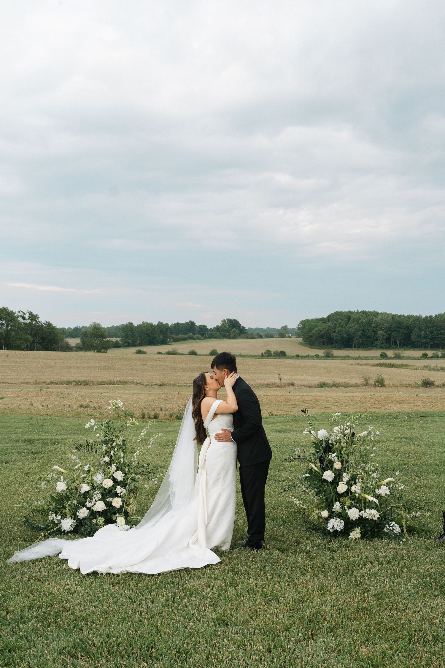 Bride in a white gown with long veil kisses groom in black suit between two white floral arrangements in a grassy field in Kentucky