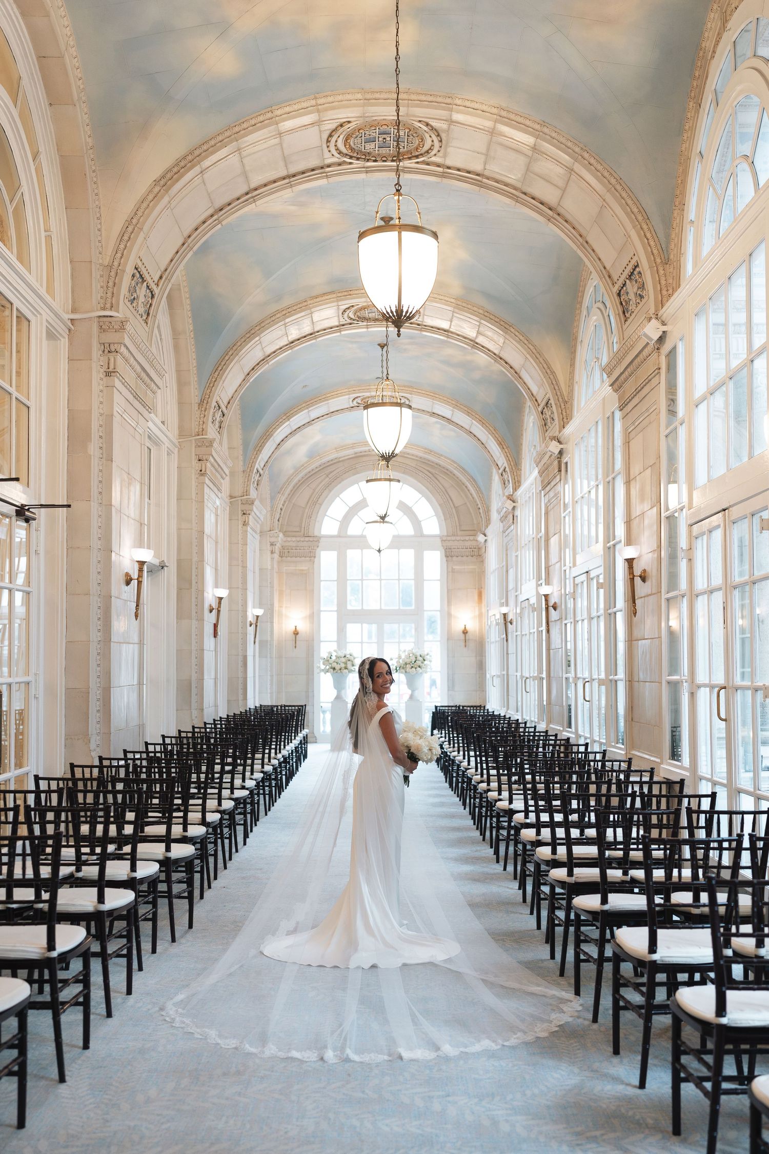 Bride in a white gown with cathedral veil stands in an elegant arched hall lined with black chairs at the Hermitage Hotel in Nashville