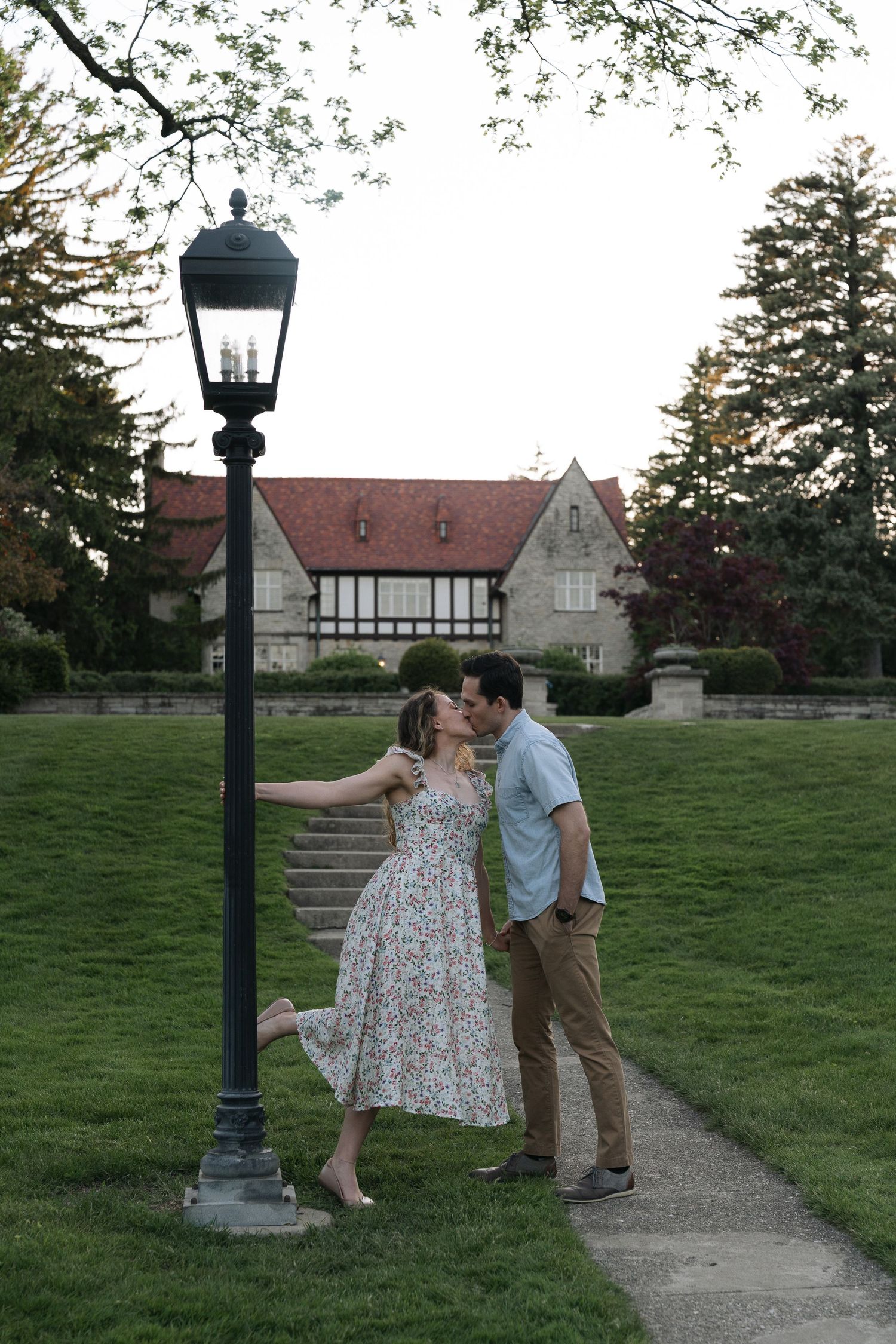 Couple kissing by a vintage lamppost on a garden path with a Tudor-style mansion in the background.