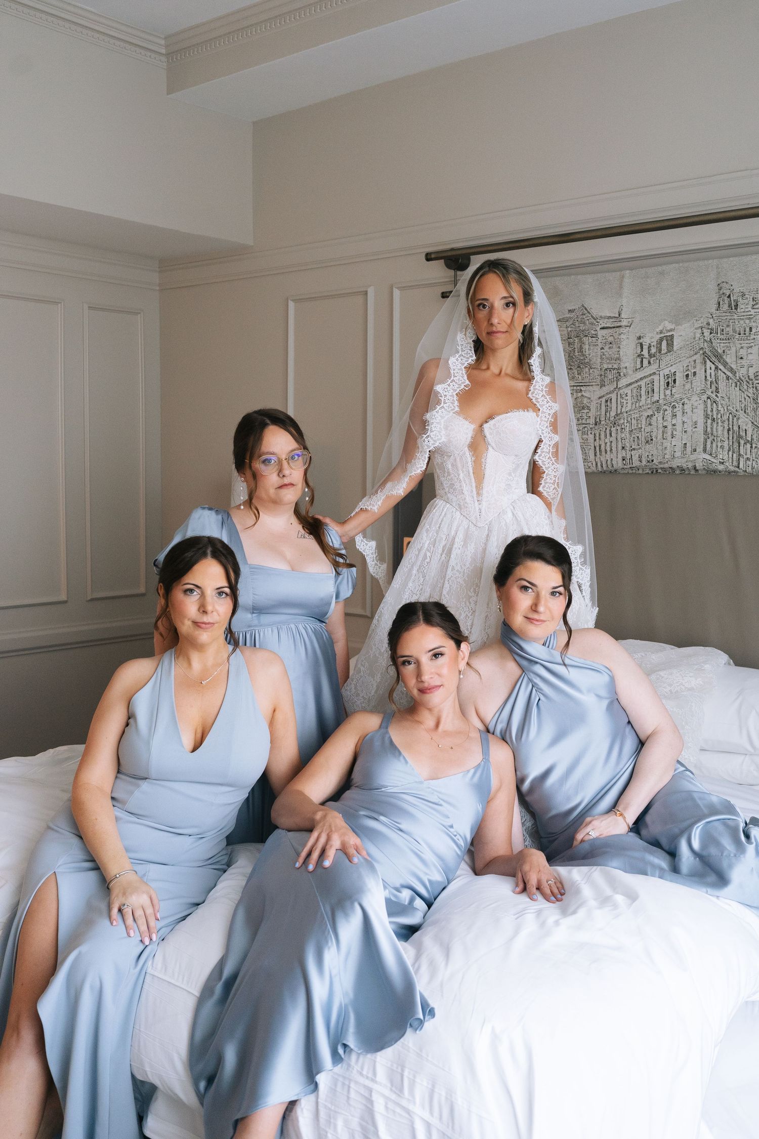 Bride in white gown and veil stands behind four bridesmaids in blue satin dresses seated on a bed.