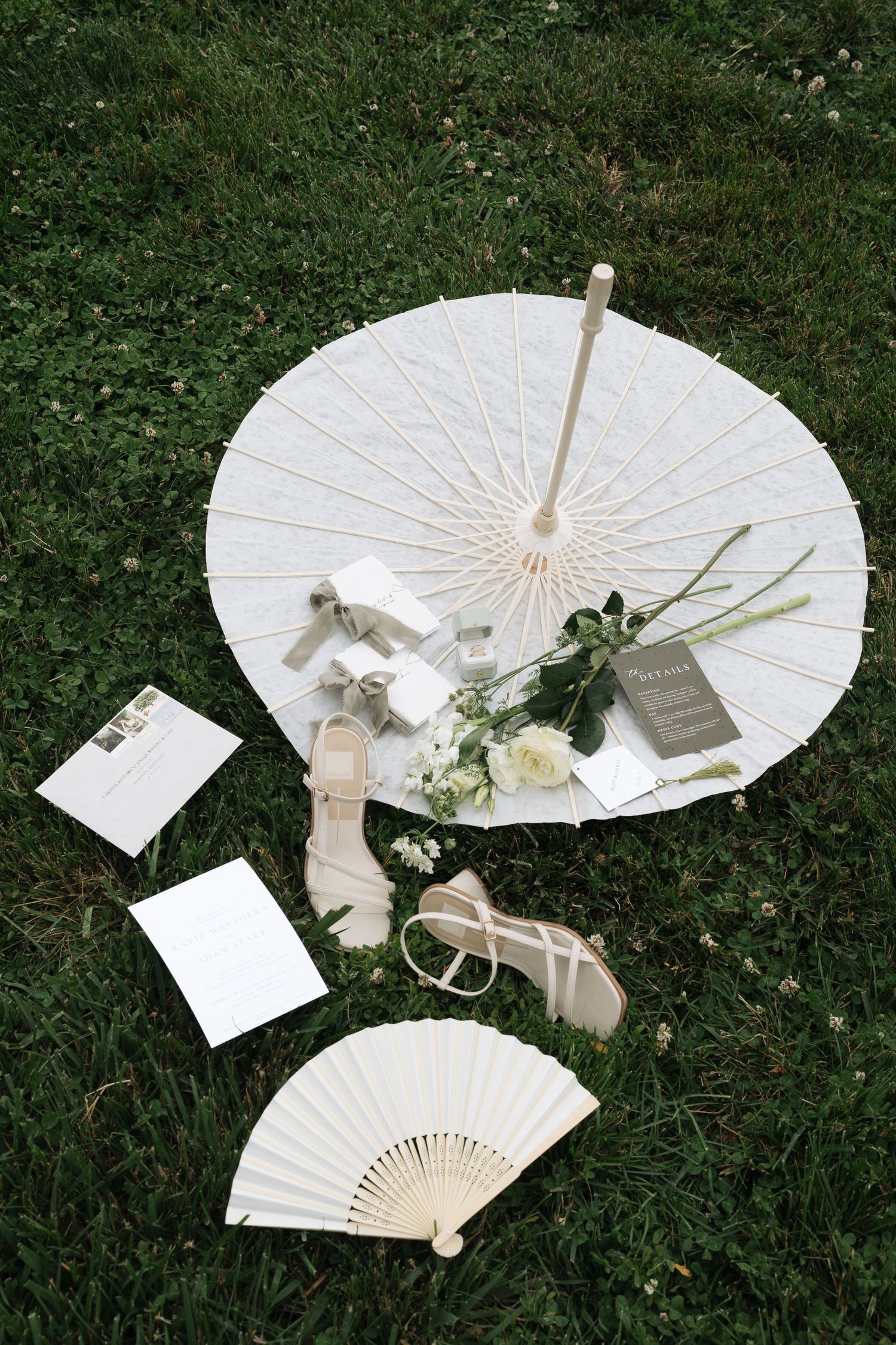 White paper parasol, folding fan, strappy sandals, bouquet and wedding invitations arranged on green grass in Kentucky