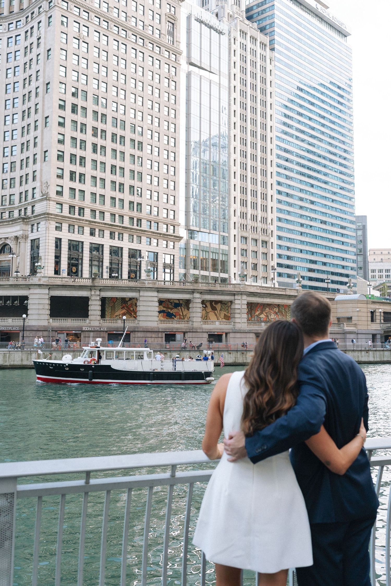 A couple overlooks the Chicago River with architectural landmarks and a tour boat passing beneath skyscrapers.