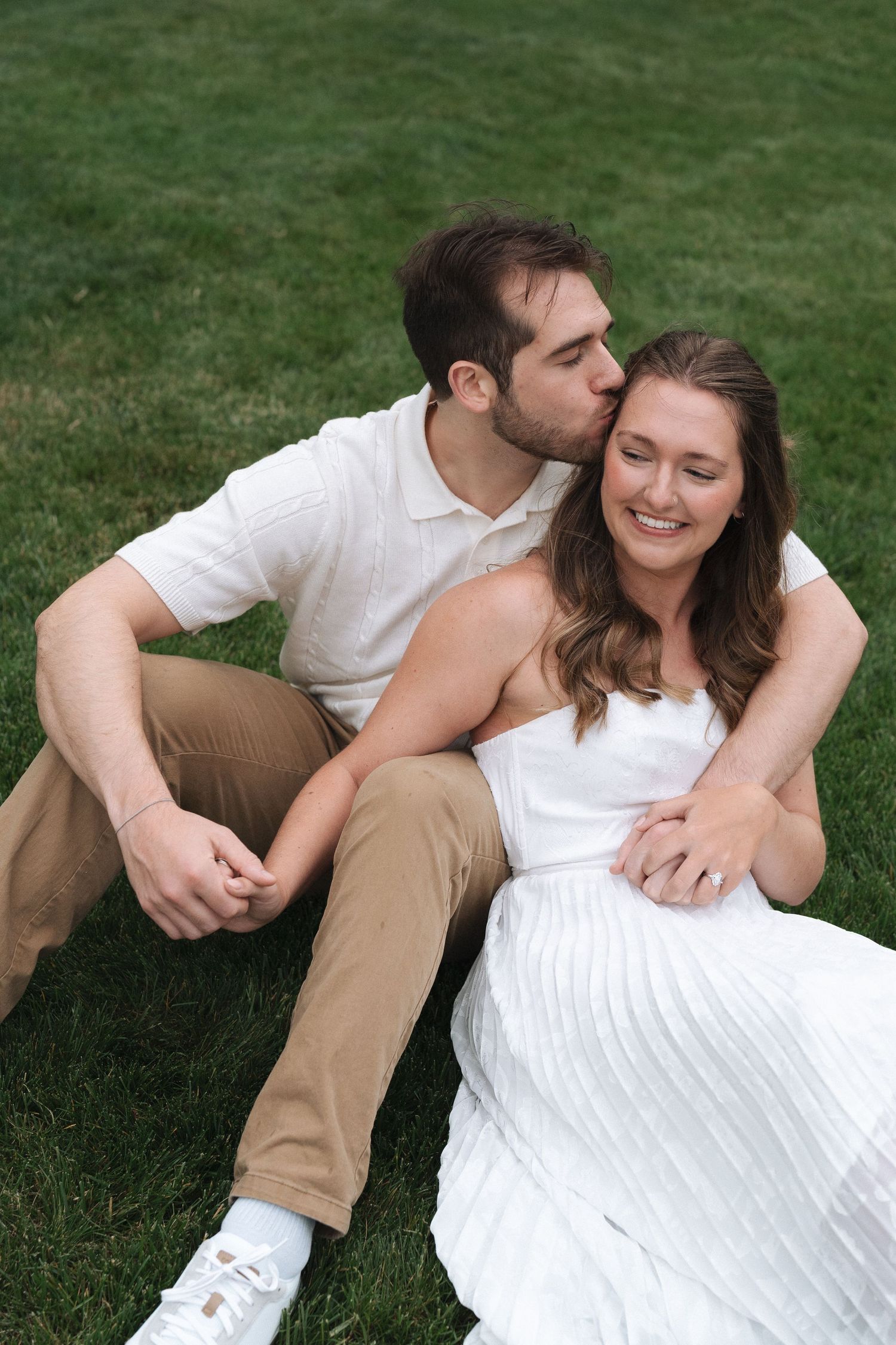 A romantic couple poses together on green grass while wearing white and tan outfits in a playful outdoor photo sequence in Carmel