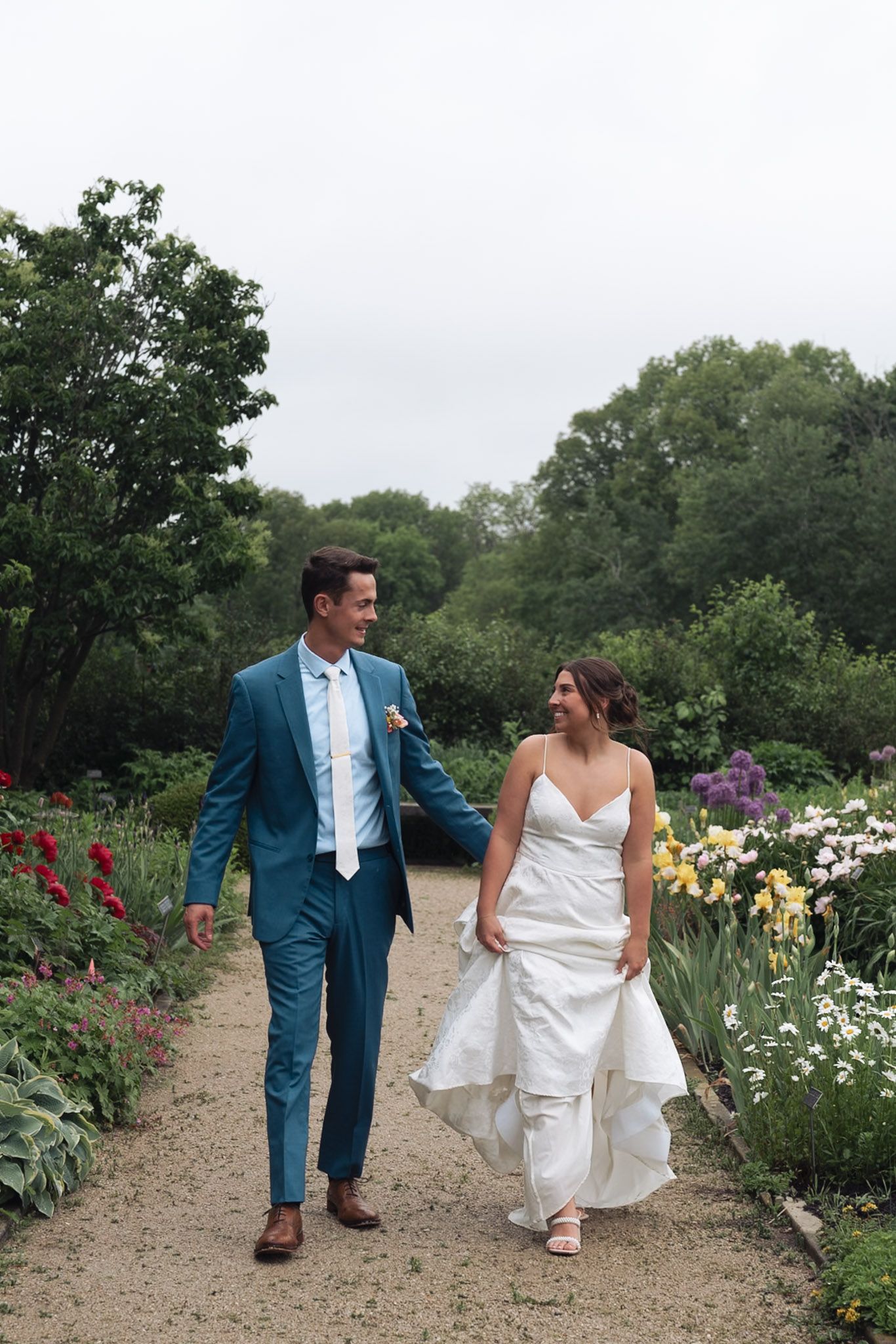 Bride and groom walk hand-in-hand along a garden path lined with colorful flowers, groom in blue suit, bride in white gown.