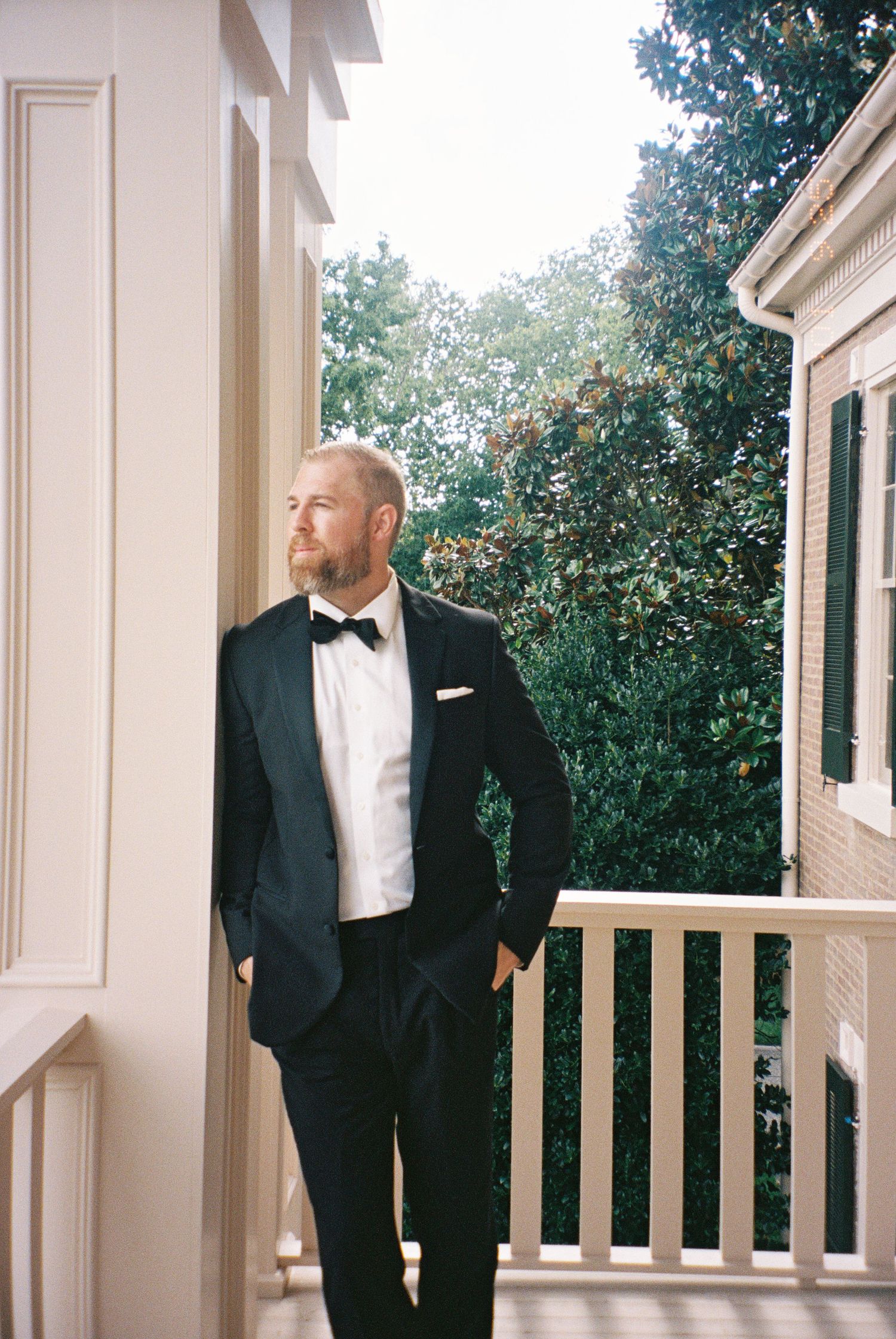 Groom in black tuxedo stand on white columned porch during elegant wedding photoshoot in Nashville