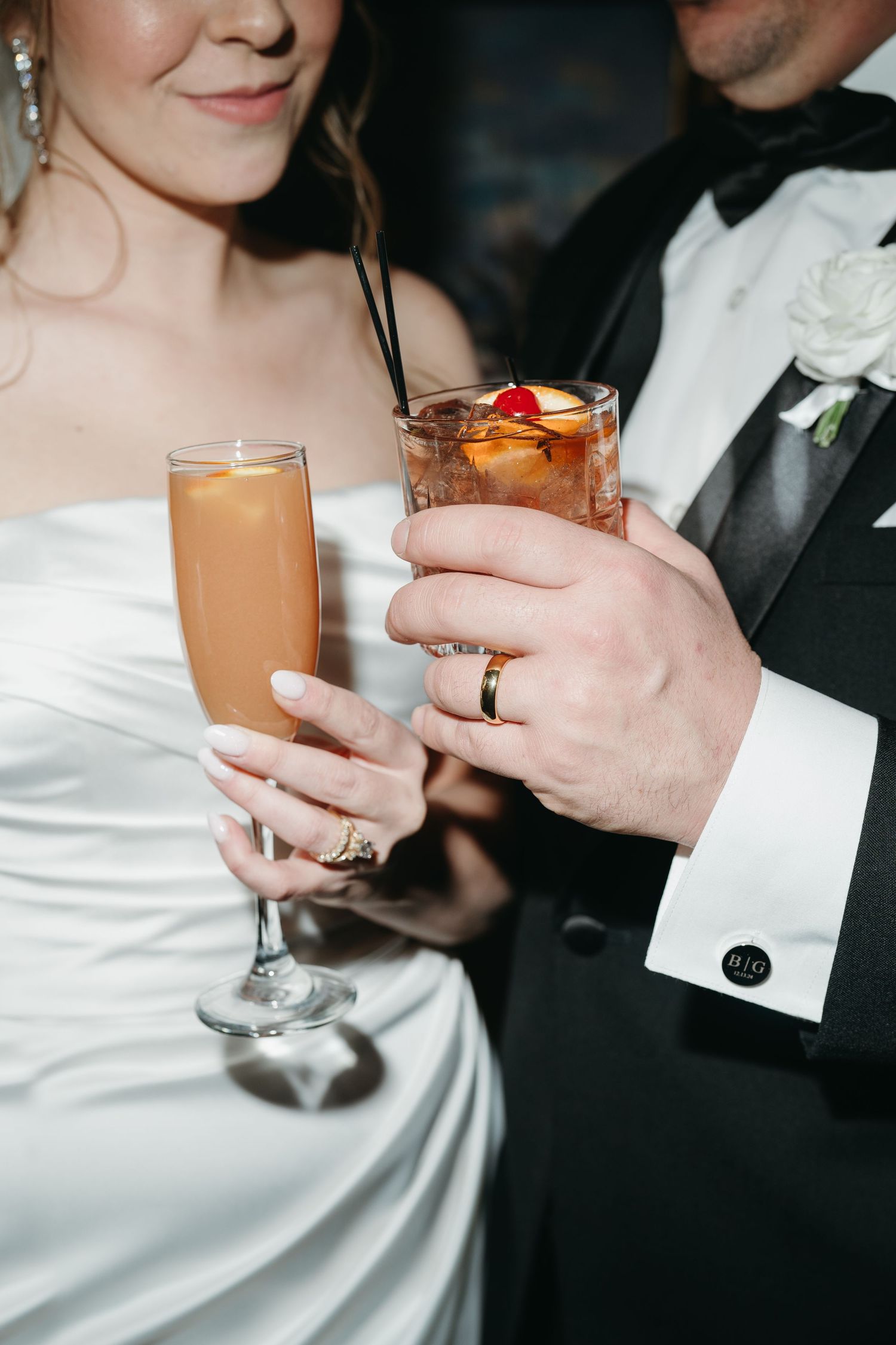 Wedding couple holds cocktail glasses while displaying elegant wedding rings at reception celebration.