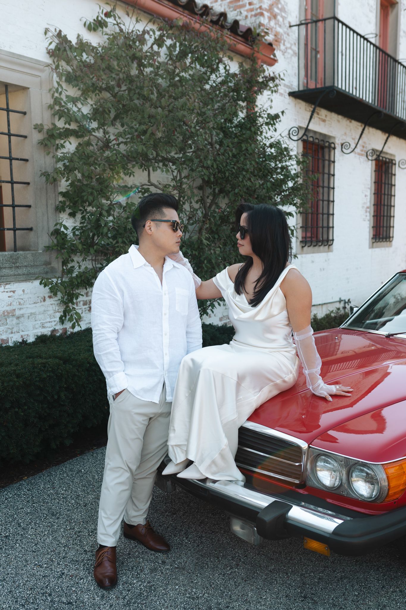 A couple in white outfits poses romantically against a vintage red sports car outside a Spanish-style building.