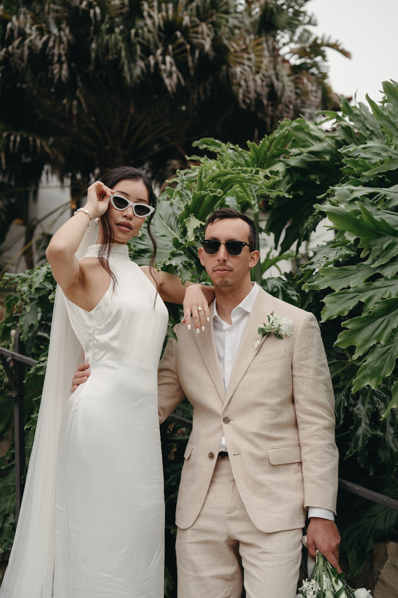 A couple in a white dress and beige suit poses intimately together among tropical foliage during their wedding photoshoot in Santa Barbara