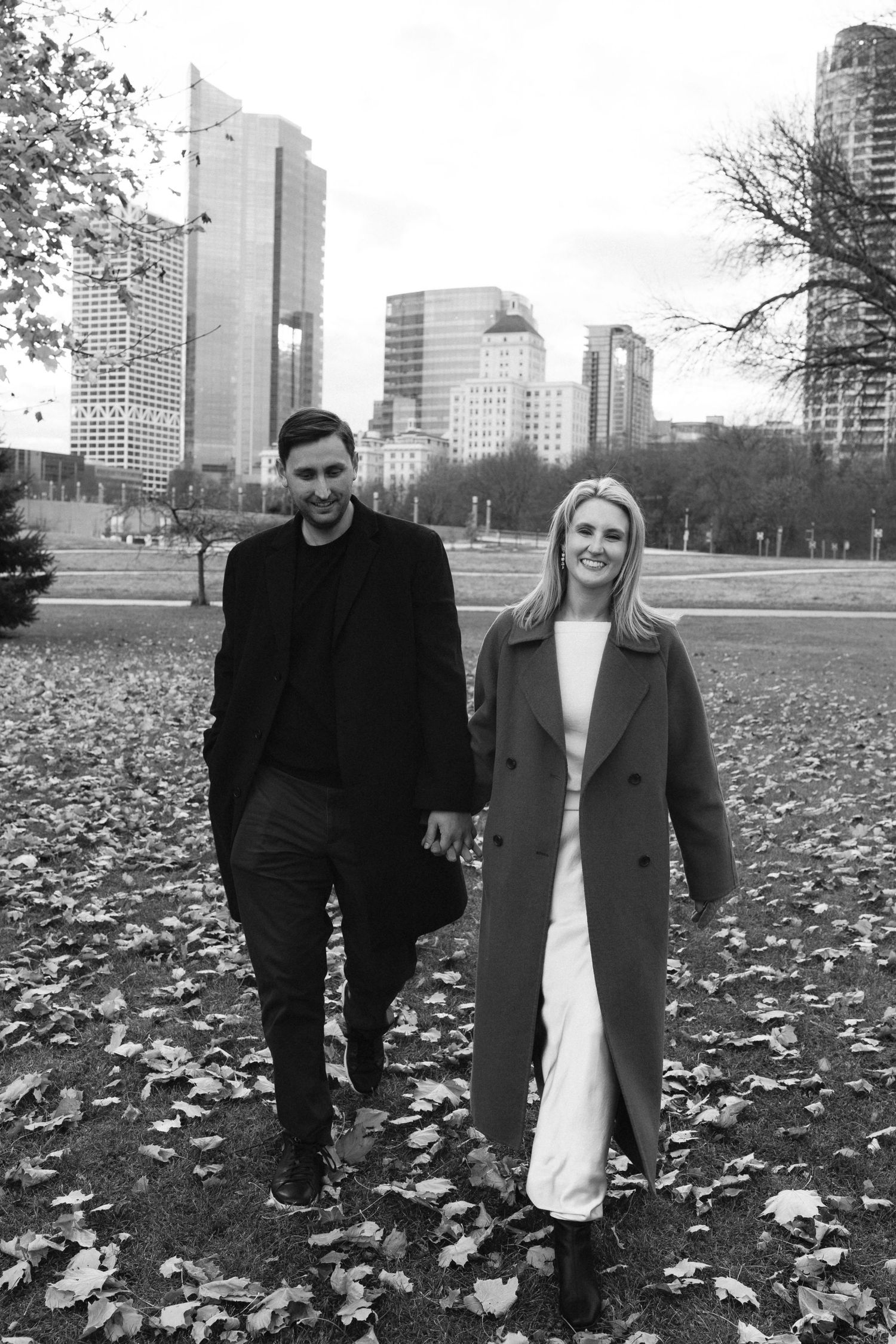 A couple holds hands walking through a leaf-covered park with city skyscrapers in the background on a fall day.