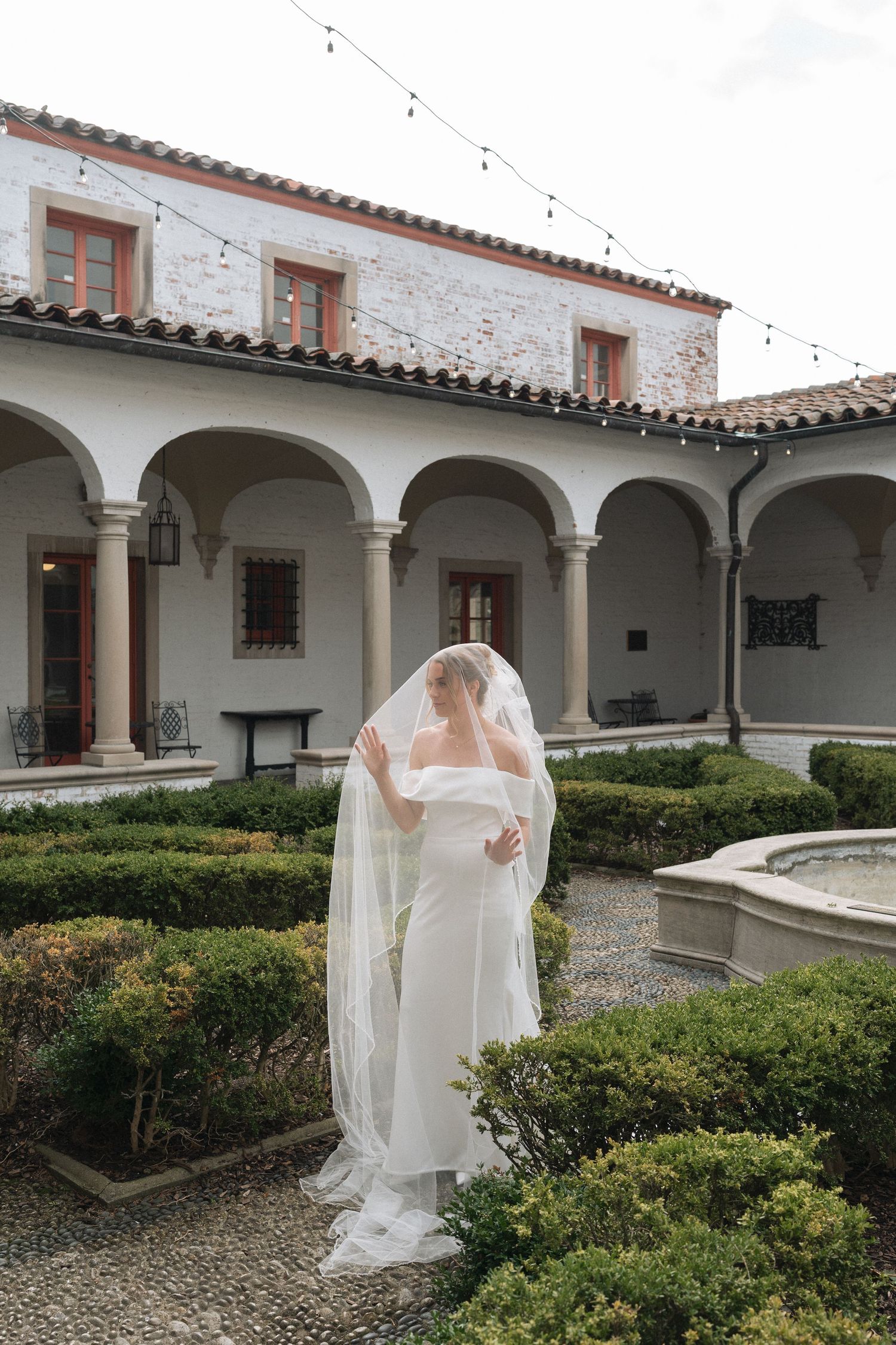 Bride in white gown and veil stands in elegant Italian courtyard garden with arched colonnade and fountain.