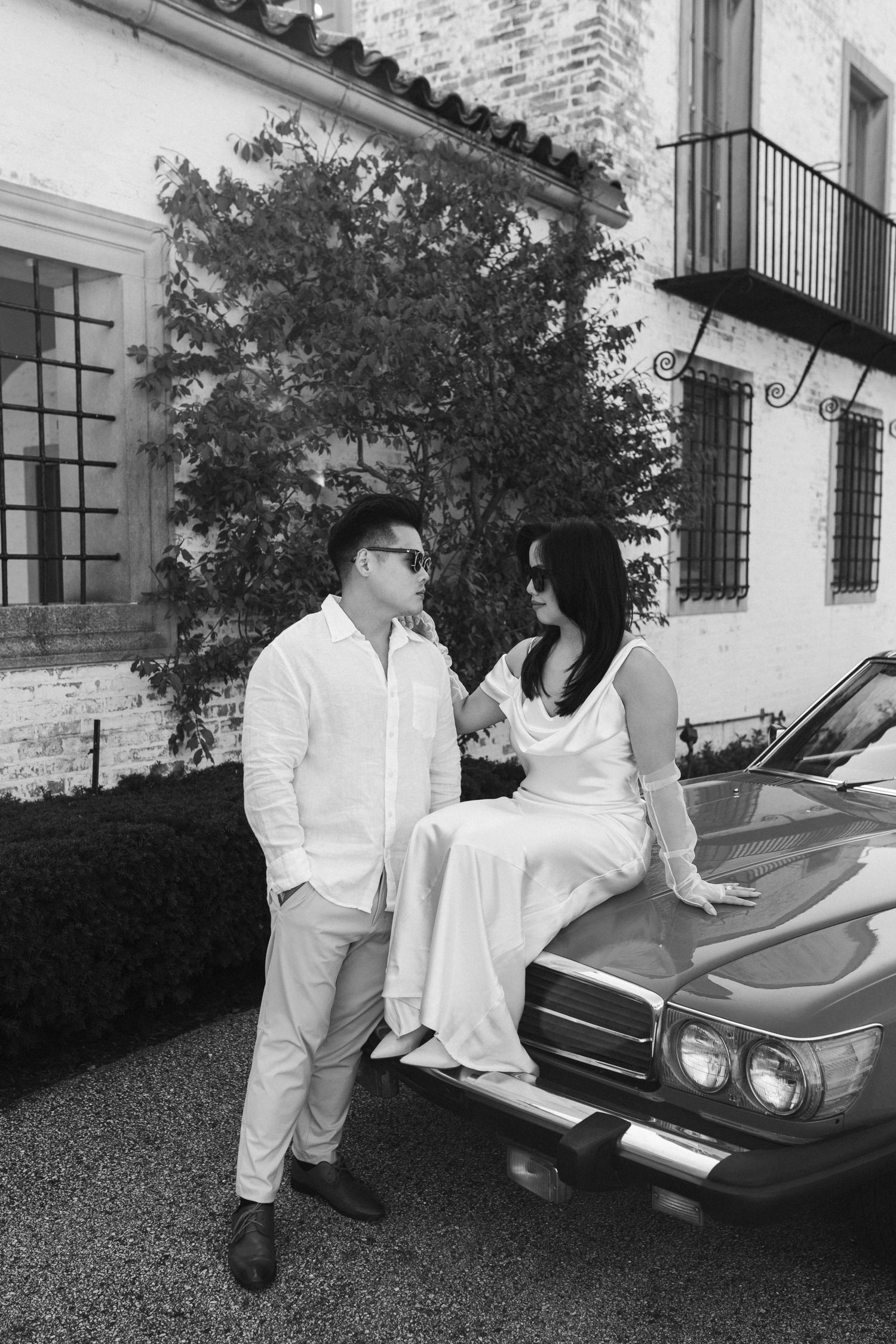 In black and white, a couple poses elegantly by a vintage car outside a classic building with a romantic ambiance.