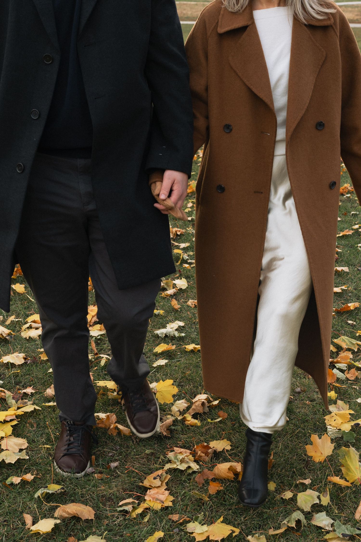 A close-up view of contrasting outfits with brown coat and white dress against black pants on autumn leaves.